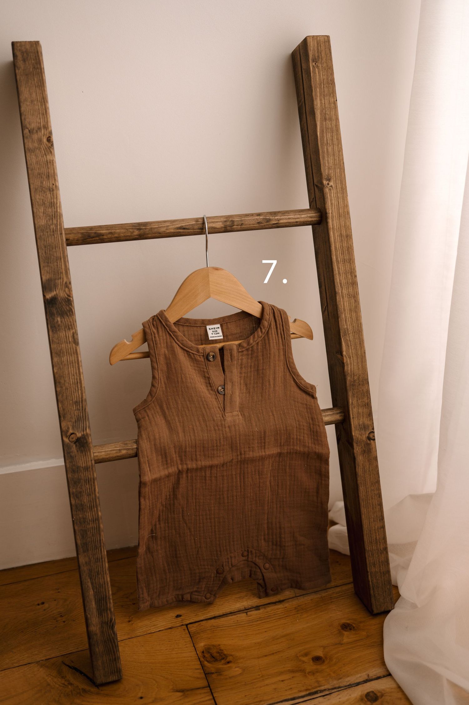 Vintage-style cotton romper in earthy brown hanging on wooden display ladder with white backdrop.