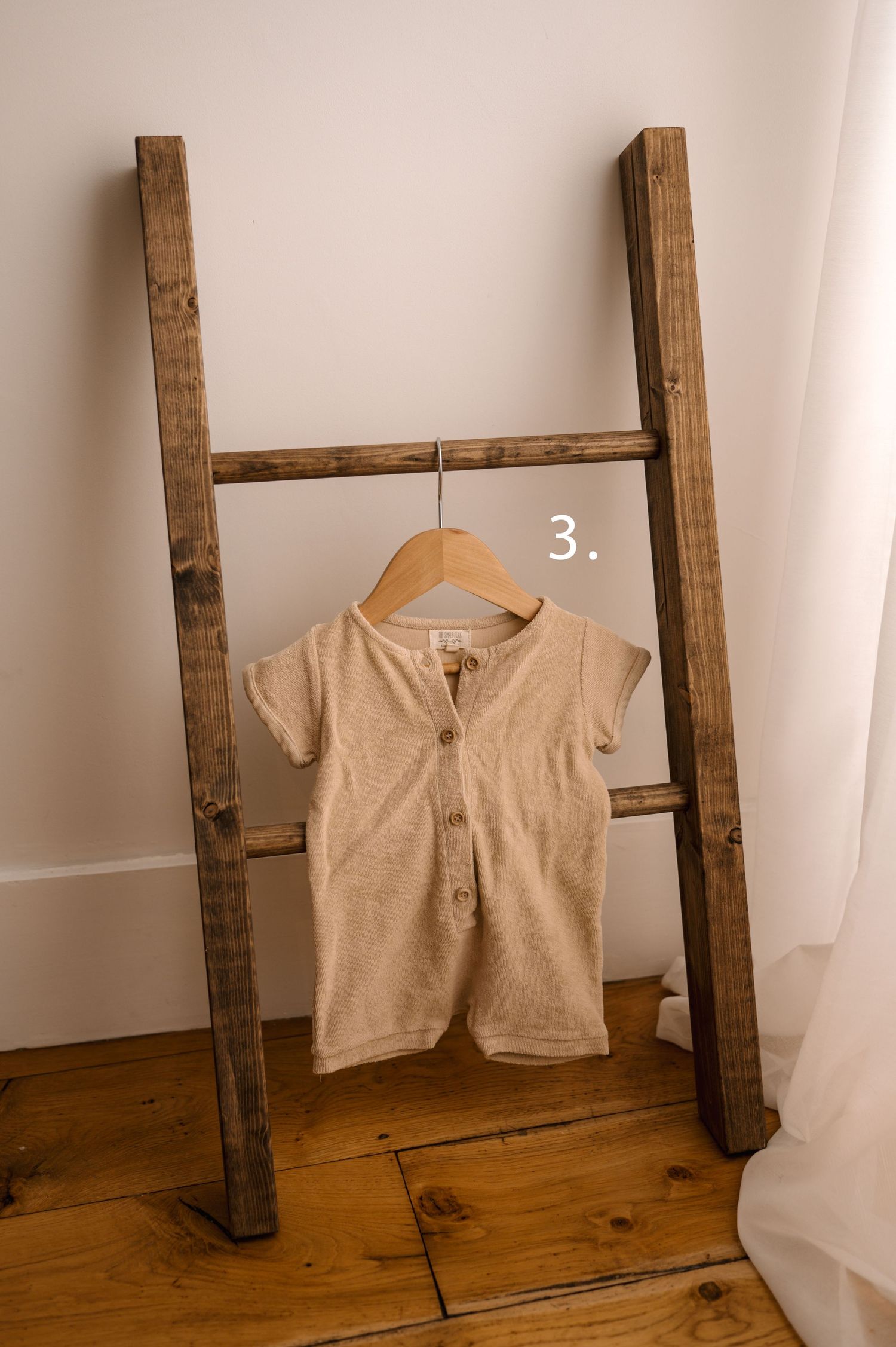 A beige short-sleeve henley shirt hangs on a wooden hanger against a rustic ladder display on hardwood flooring.