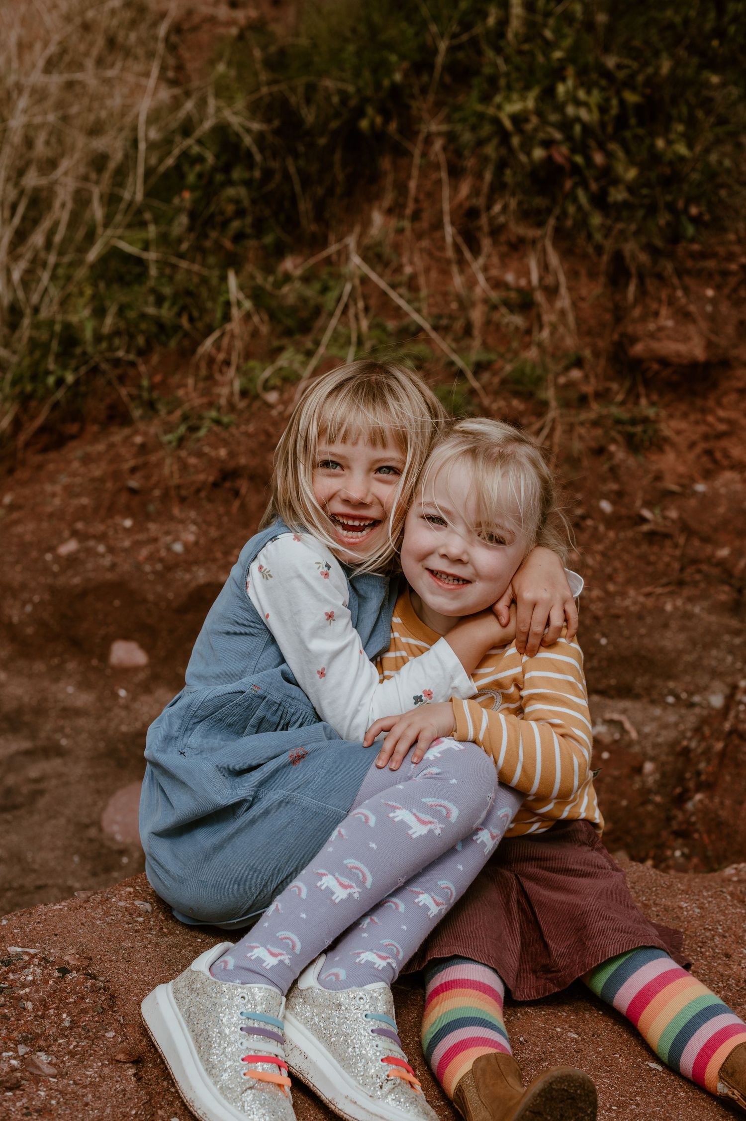 Two young children in colorful striped socks sit together and hug while laughing outdoors on a dirt path.