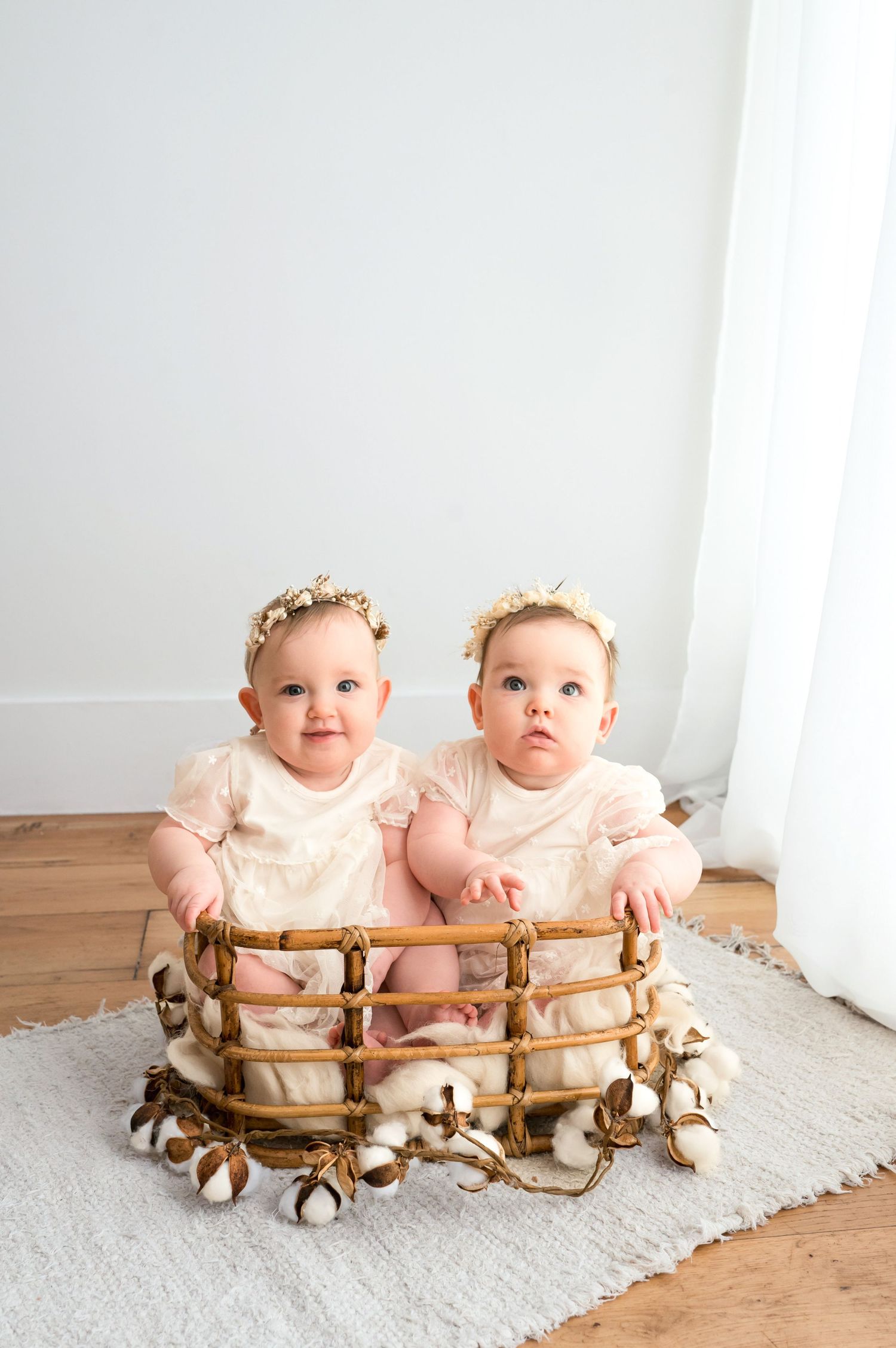 Two adorable twin baby girls in matching cream dresses and floral headbands sitting together in a wicker basket.