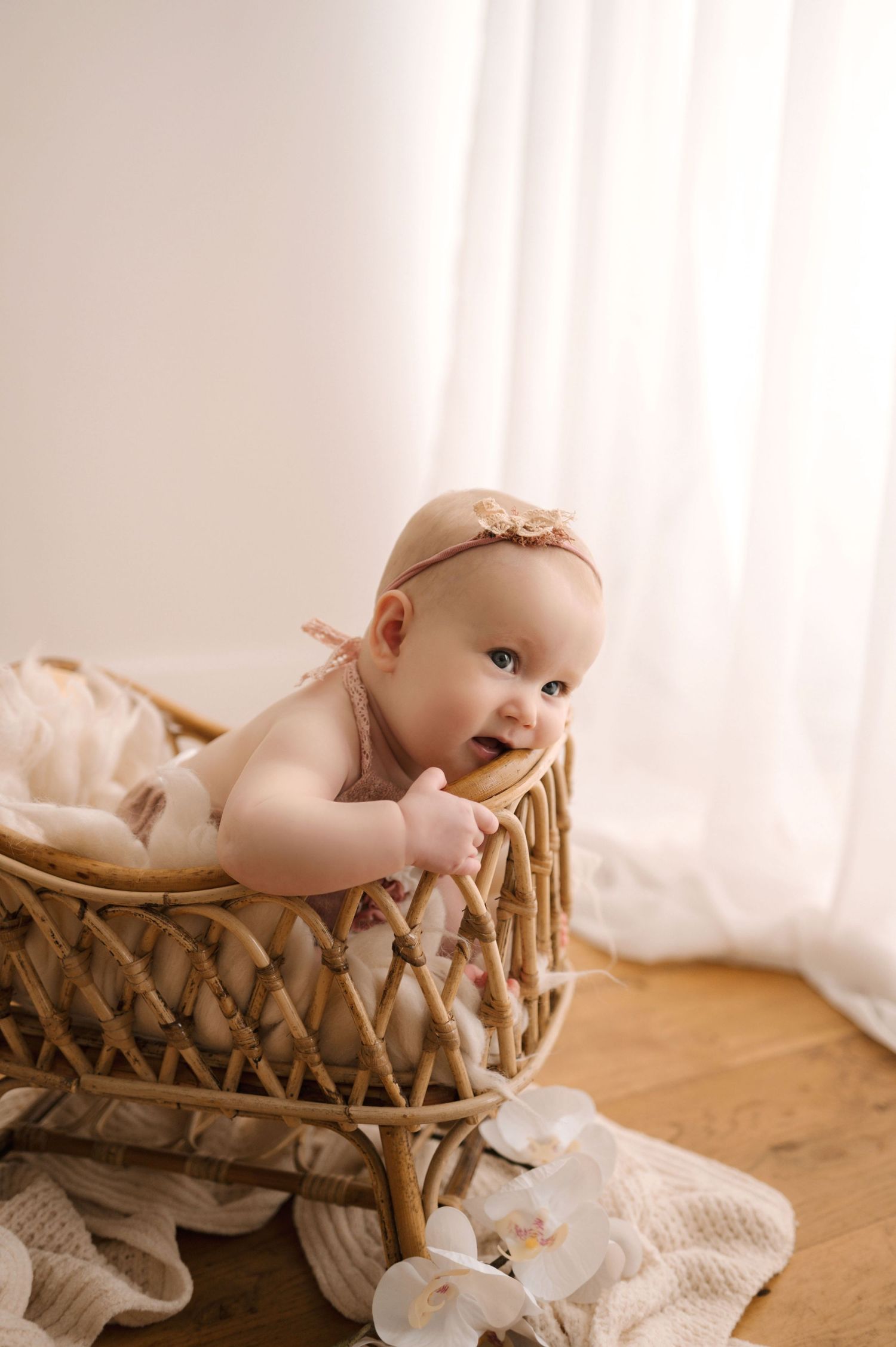 A baby girl in a floral headband peeks over the edge of a wicker bassinet lined with soft blankets.