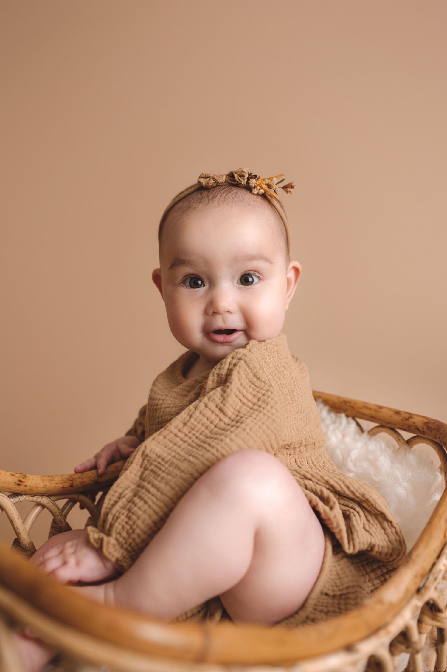 Adorable baby wearing a tan muslin outfit sits smiling in a wicker basket with white cushioning.