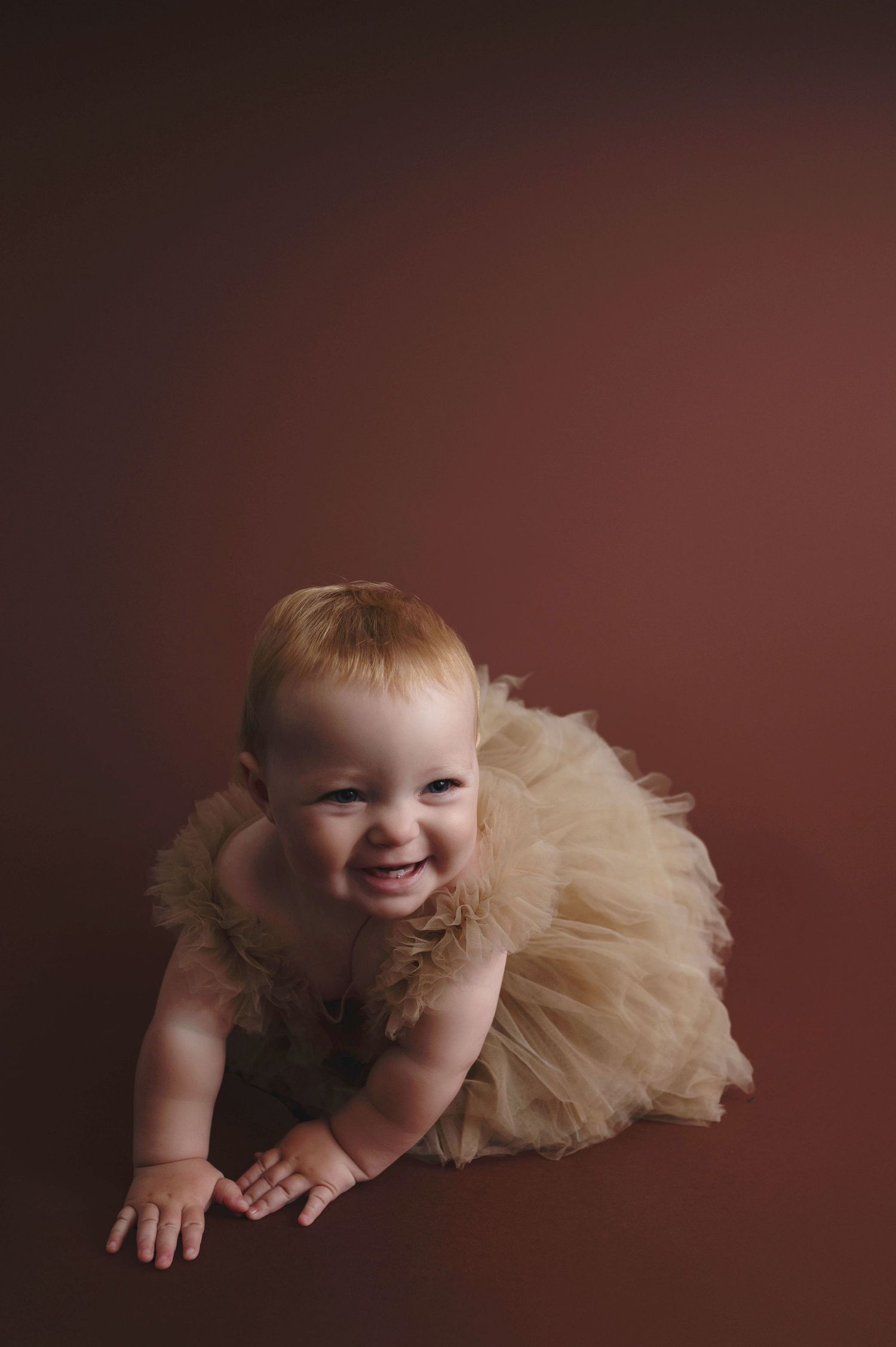Smiling baby crawling on brown backdrop while wearing a tan tulle tutu dress.