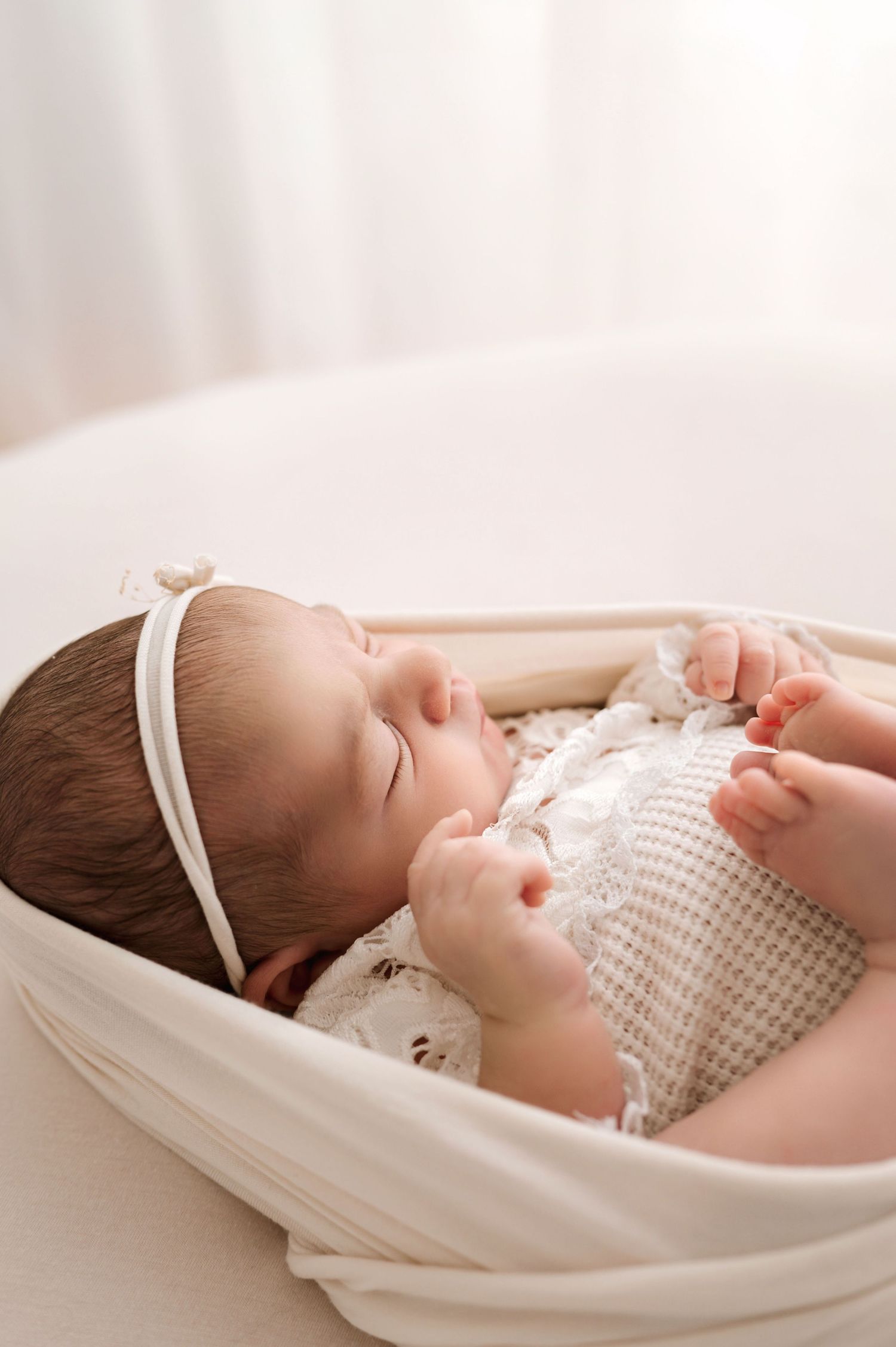 Sleeping newborn baby girl in white lace outfit and bow headband resting peacefully in a cream fabric bowl.