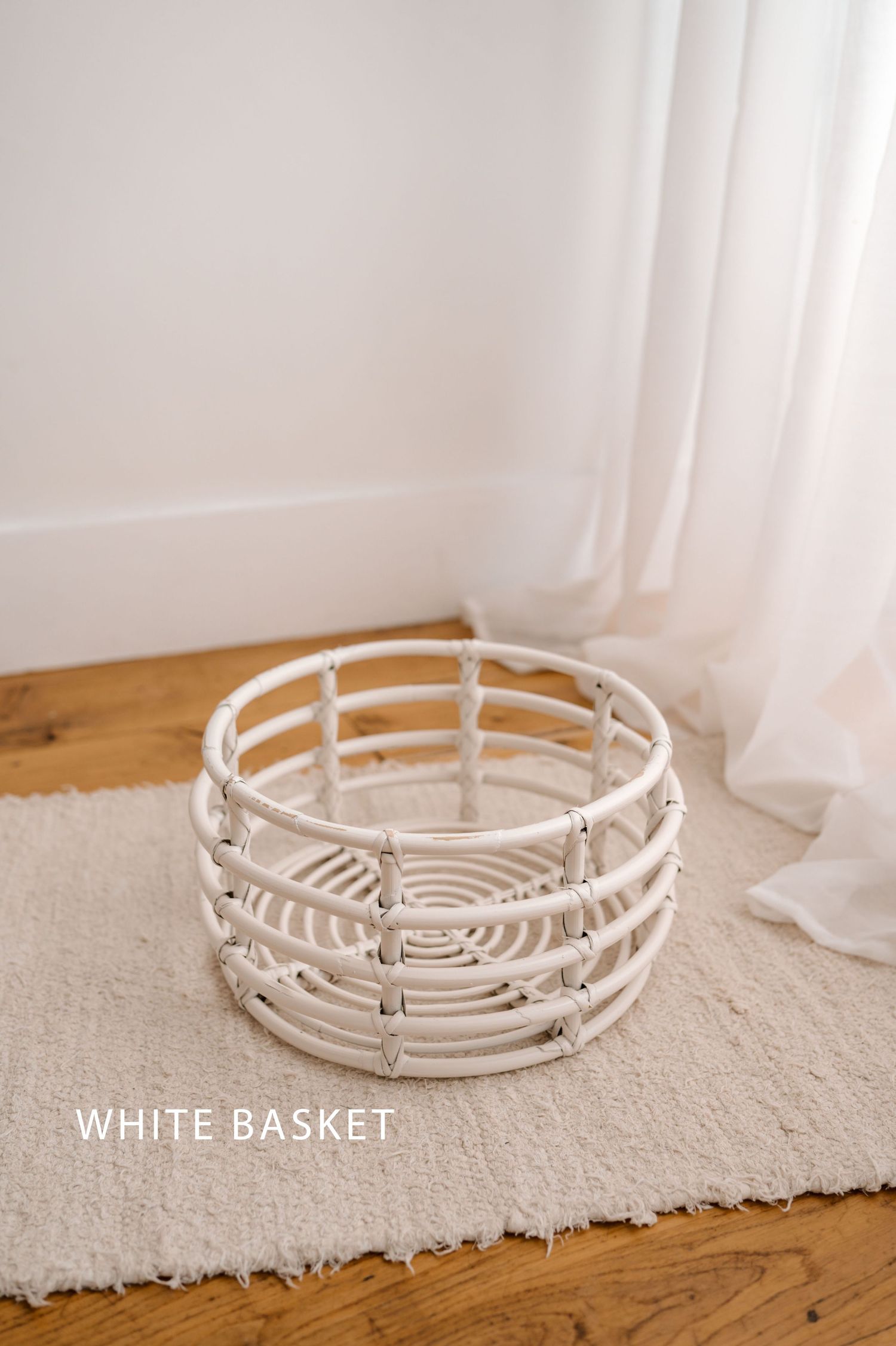 A white decorative woven basket with circular design sits on a beige carpet against wooden flooring and light curtains.