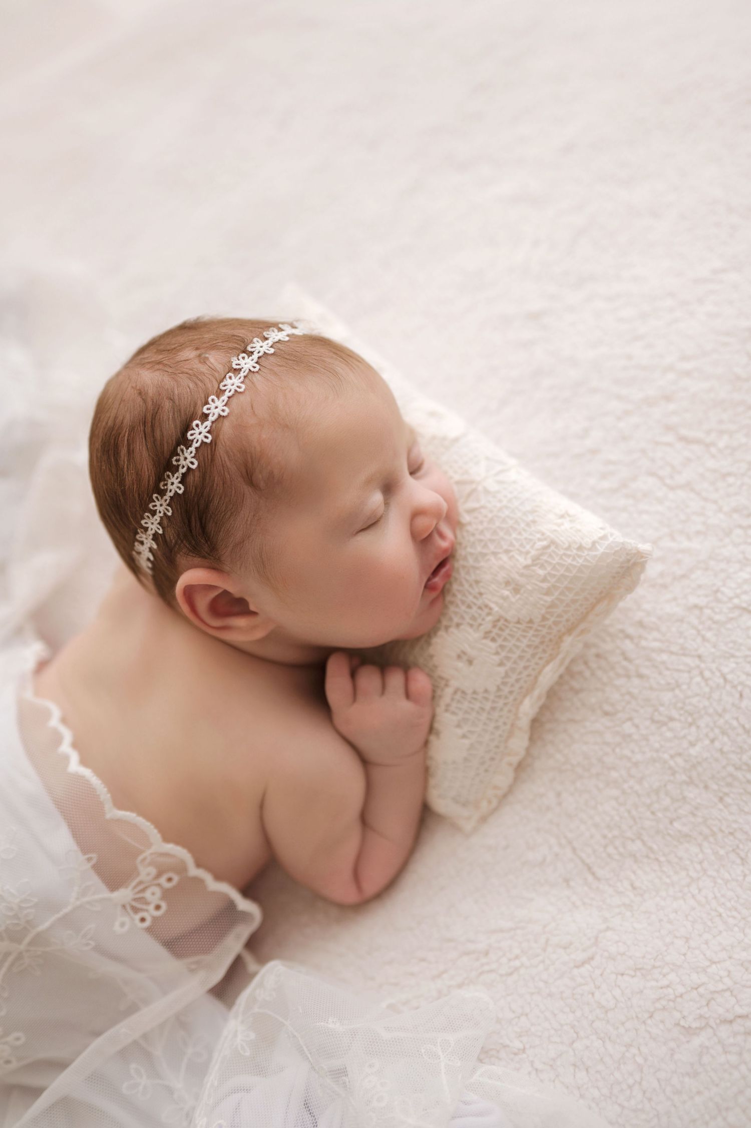 A sleeping newborn baby wearing a delicate white headband lies on soft white fabric in a peaceful pose.
