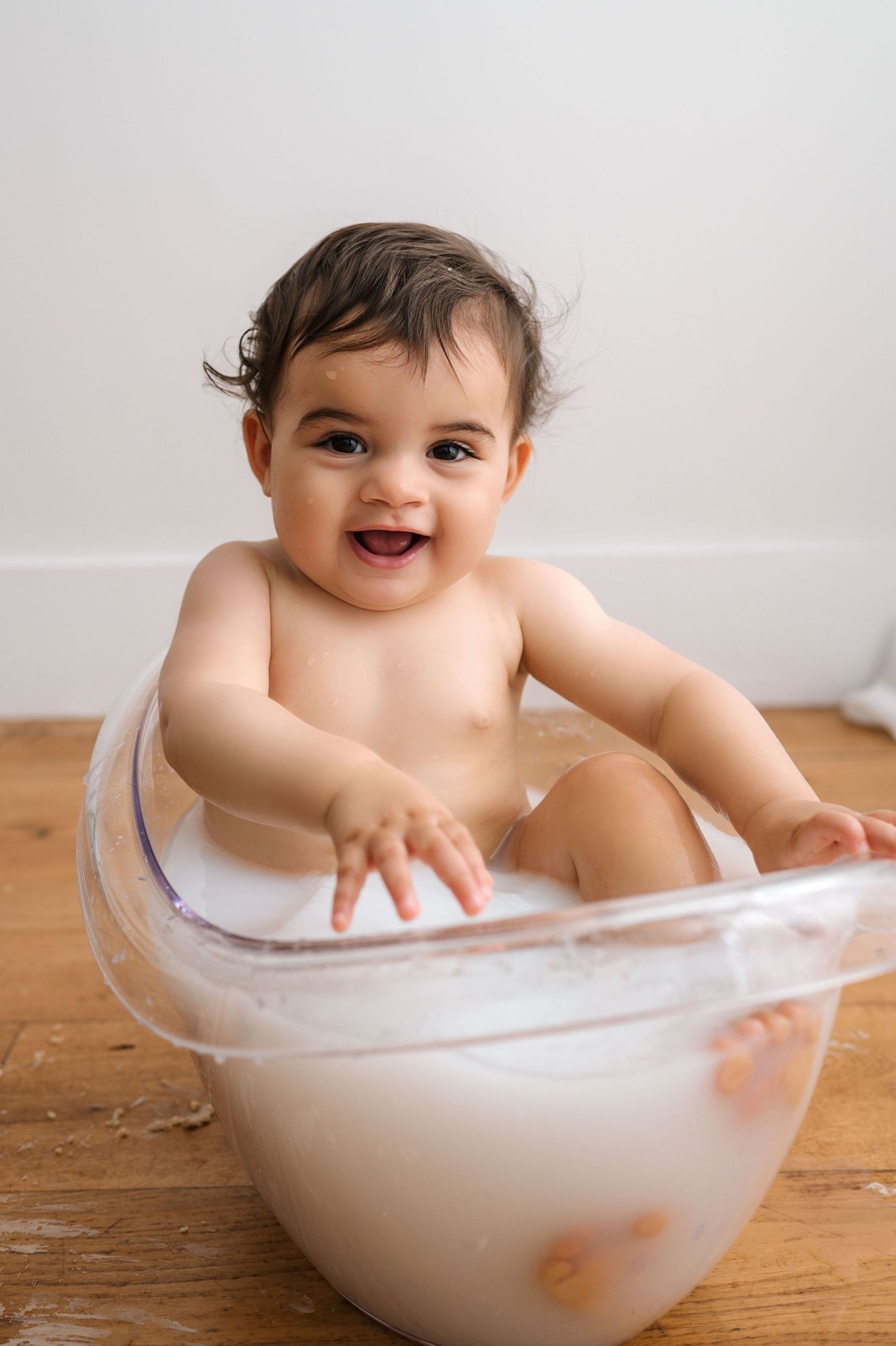 Baby sits in a white bowl with soapy bubbles during bath time on a wooden surface.