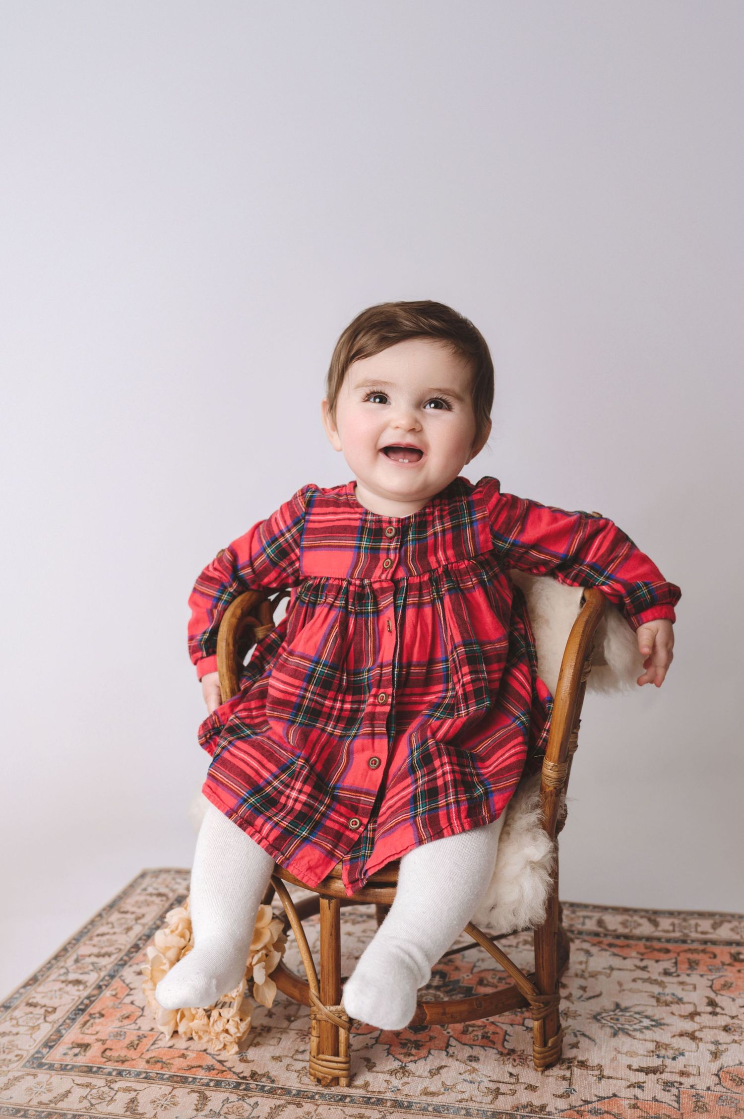 A happy baby wearing a red plaid dress and white tights sits in a wooden chair on a patterned carpet.