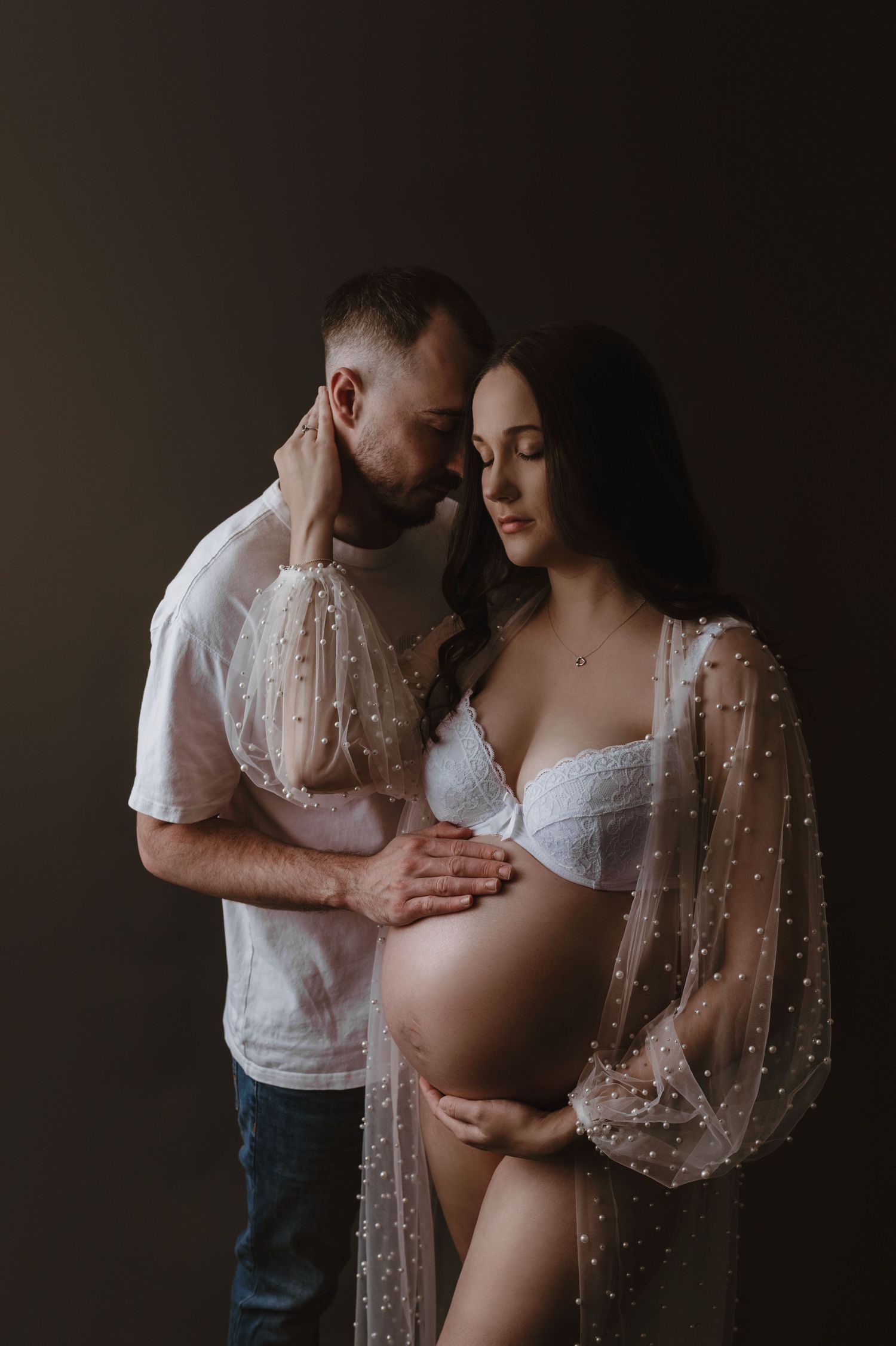 Moody maternity photo of expectant couple embracing in dark room with sheer beaded robe and white lingerie.