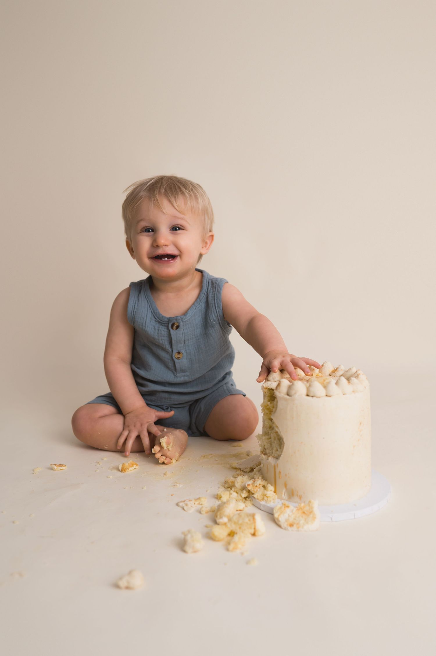 A sequence showing a baby in blue romper progressively smashing and playing with a white birthday cake during a cake smash photo shoot.