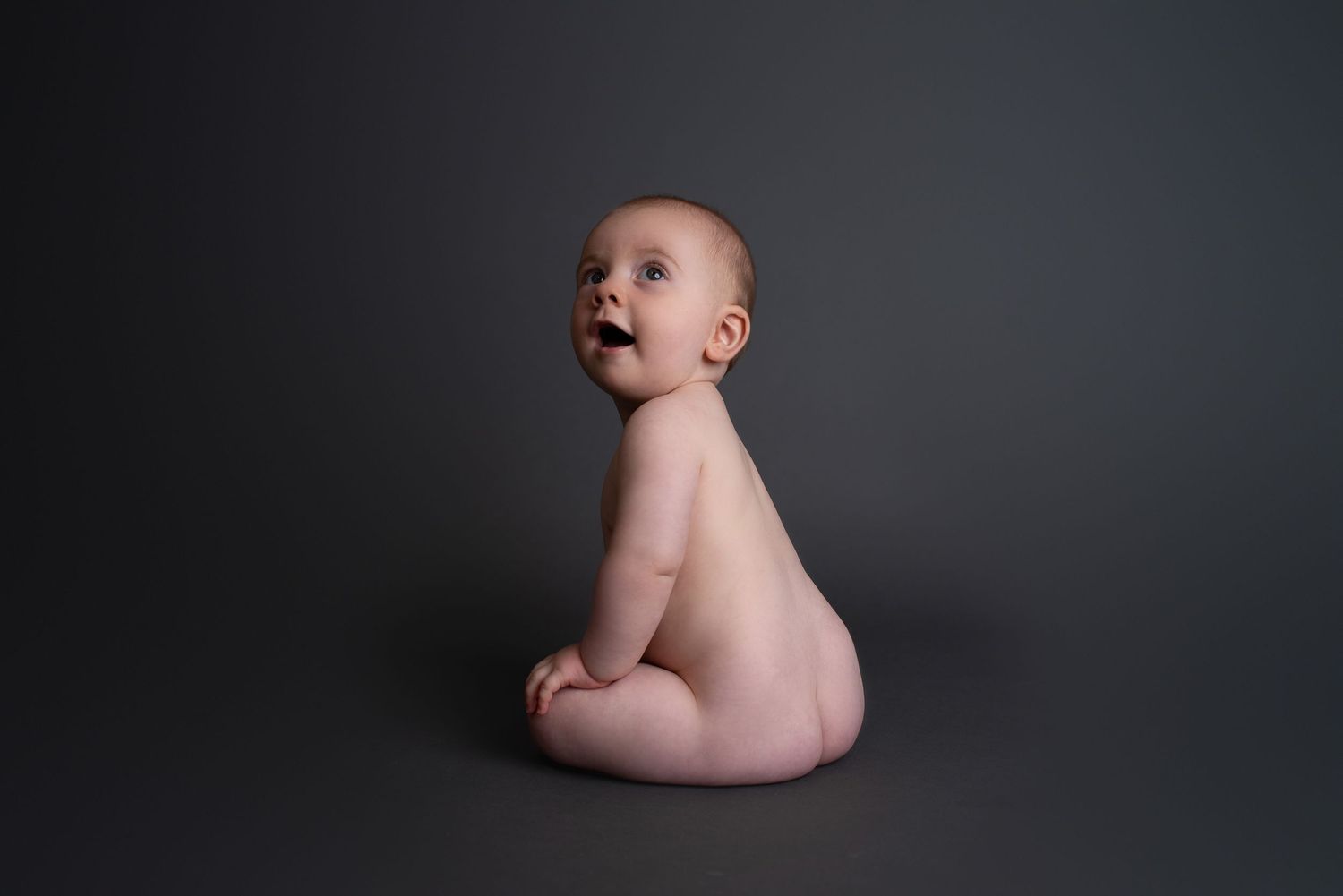 Artistic portrait of a bare baby sitting against dark background and looking upward with an expression of wonder.