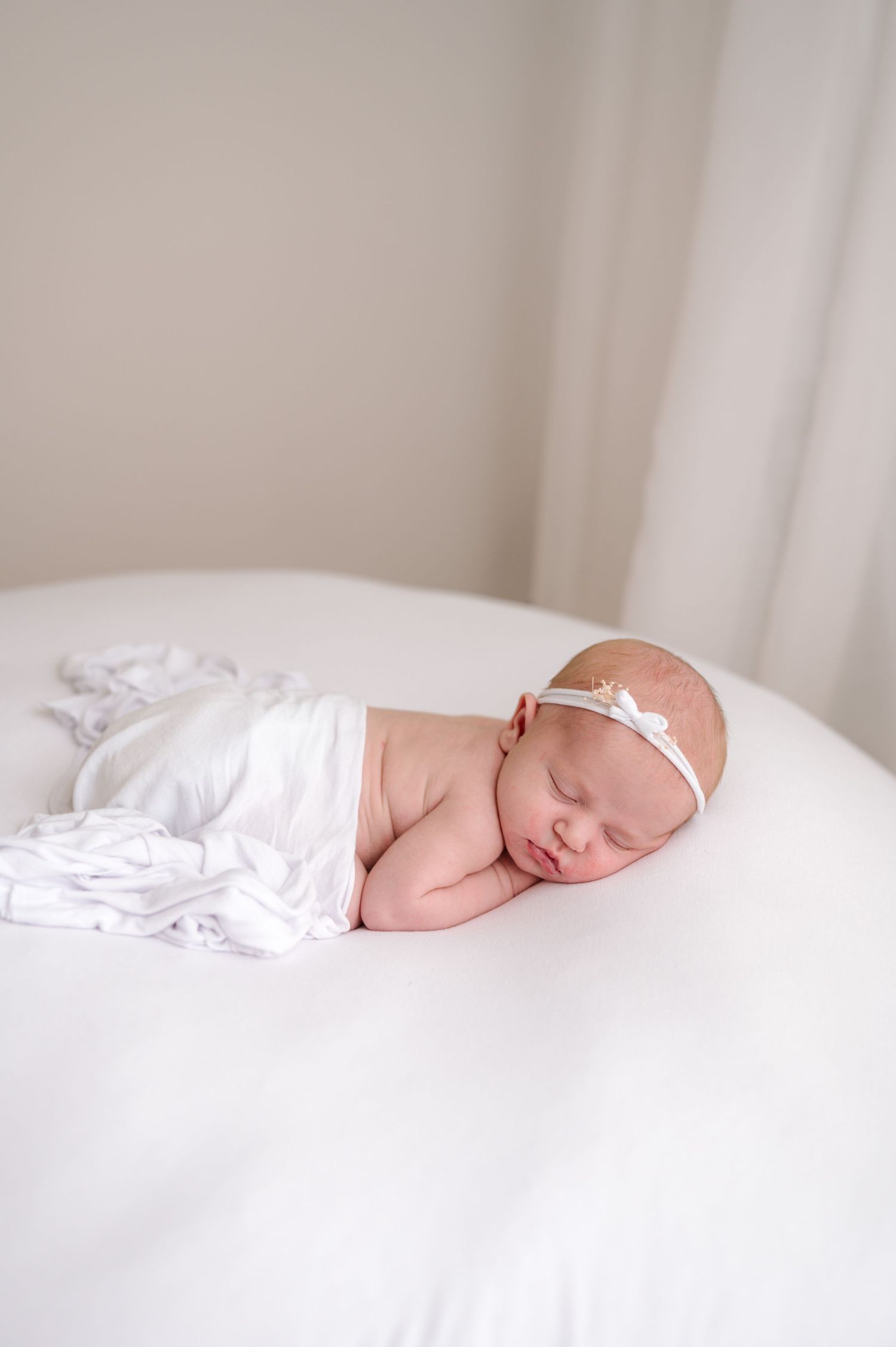 Sleeping newborn baby girl wrapped in white swaddle with delicate headband on white rounded surface.