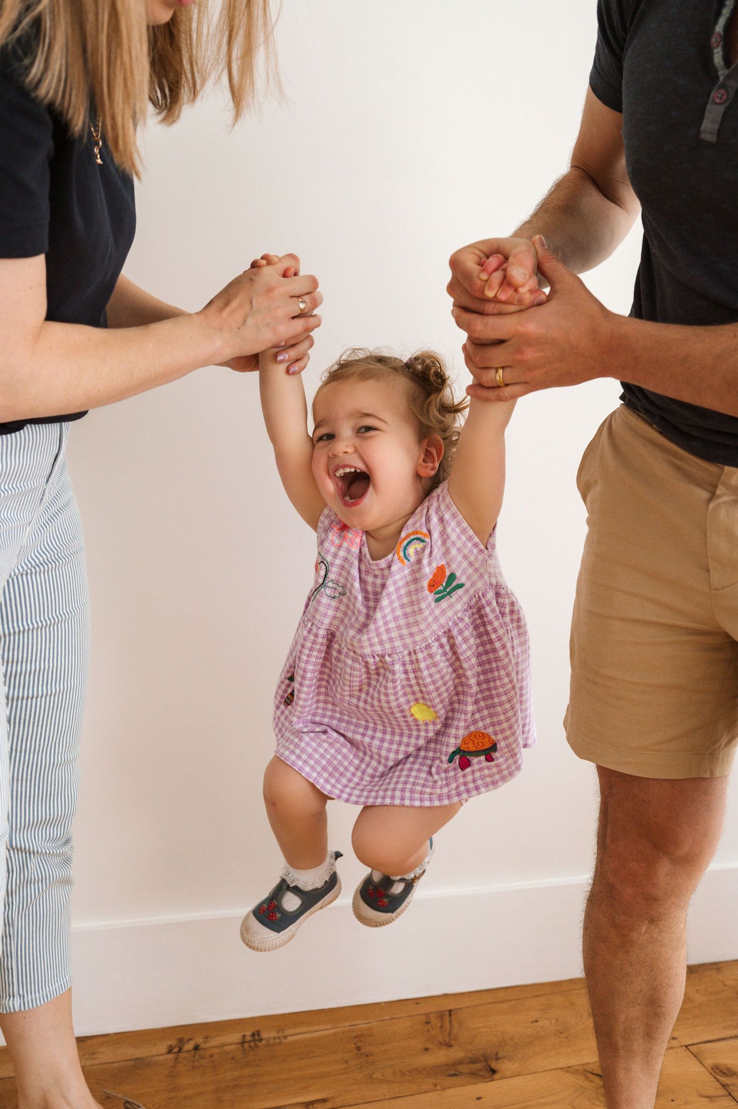 Two adults swinging laughing toddler girl in pink gingham dress between them indoors.