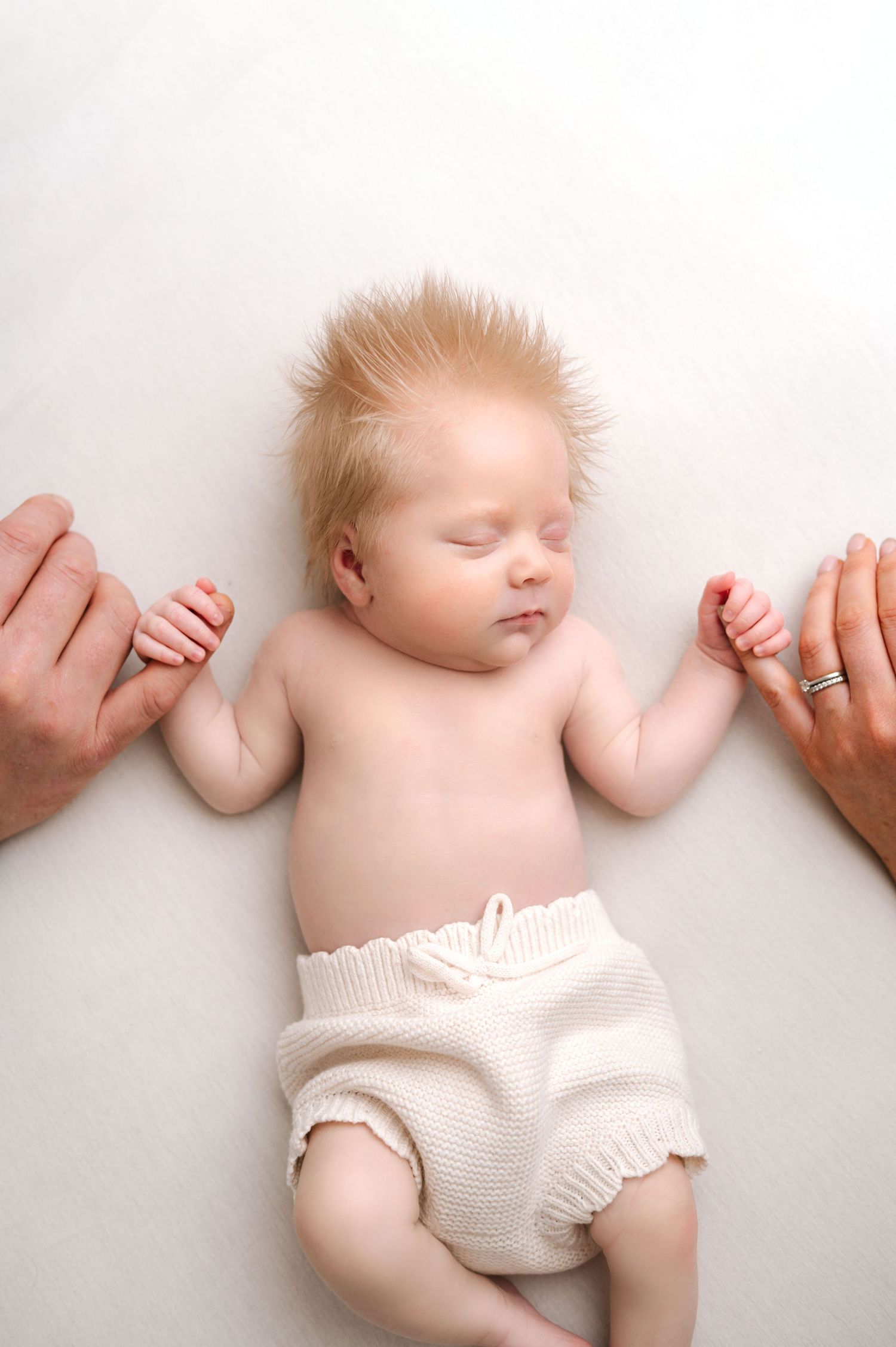 Newborn baby with blonde hair sleeping peacefully in cream knit shorts, held by parents hands on white background.