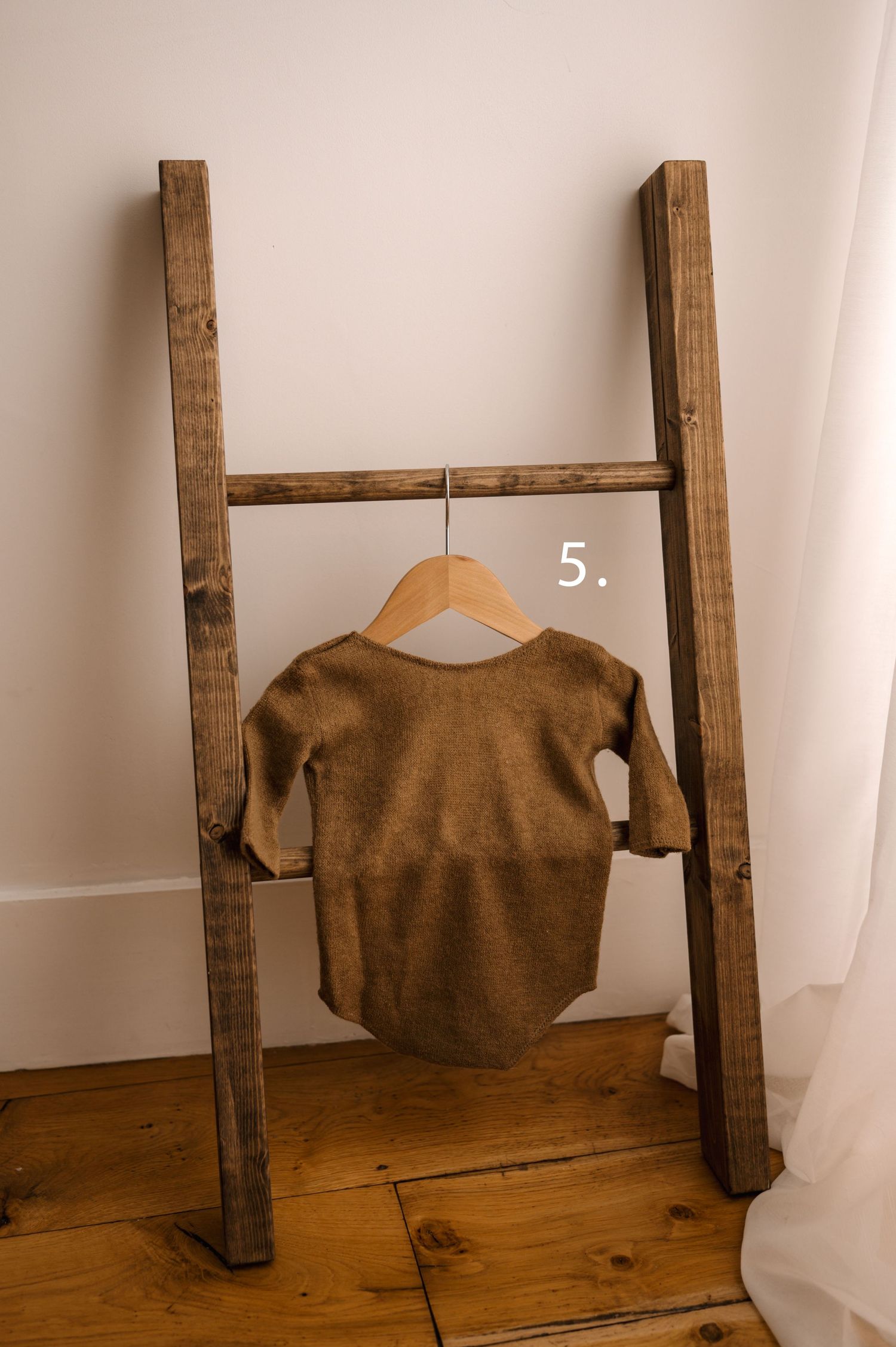 A brown baby bodysuit displayed on a wooden clothes hanger leaning against a rustic ladder.