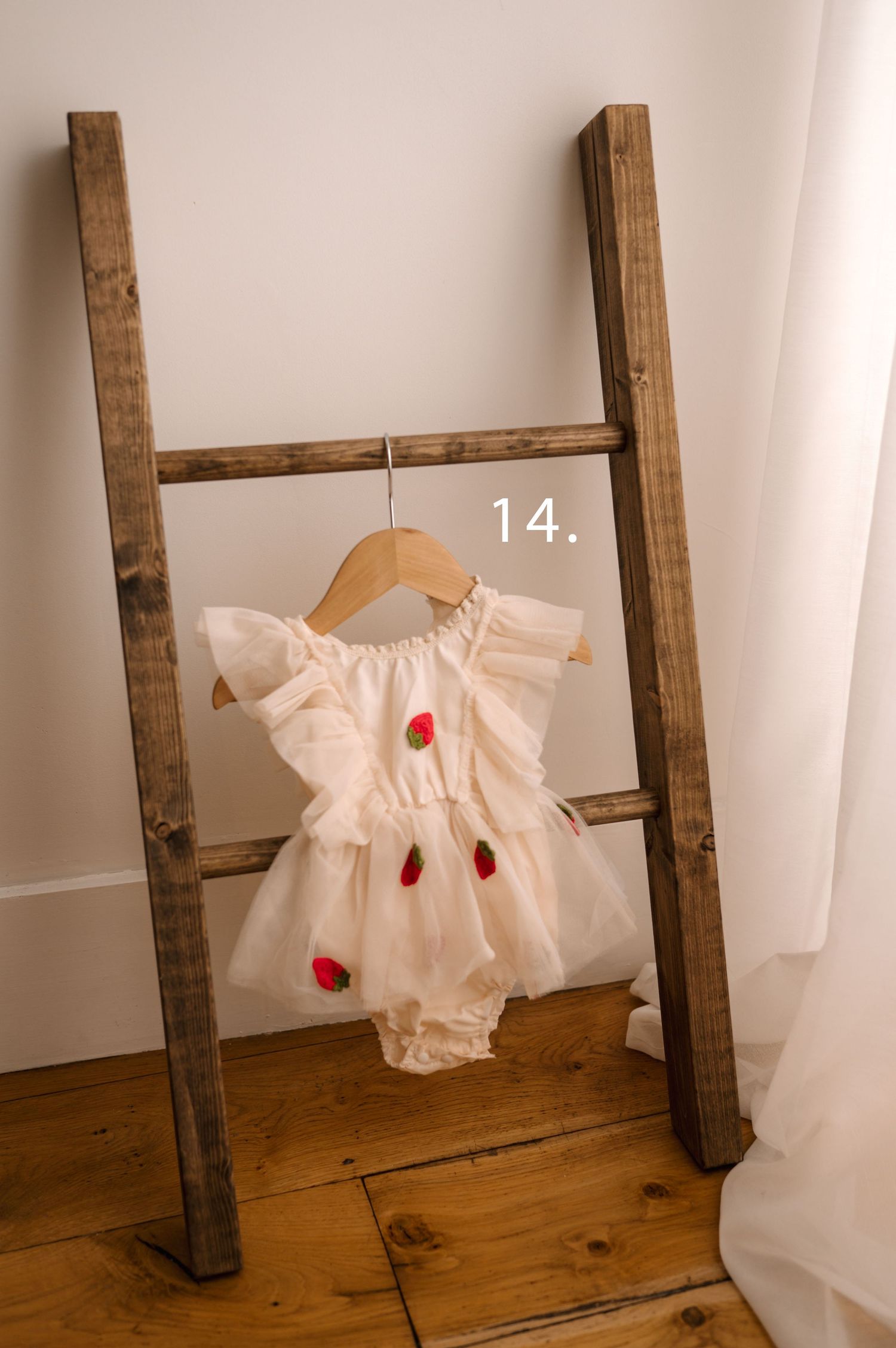 A delicate white baby dress with red strawberry embroidery hangs on a wooden hanger against a rustic ladder backdrop.