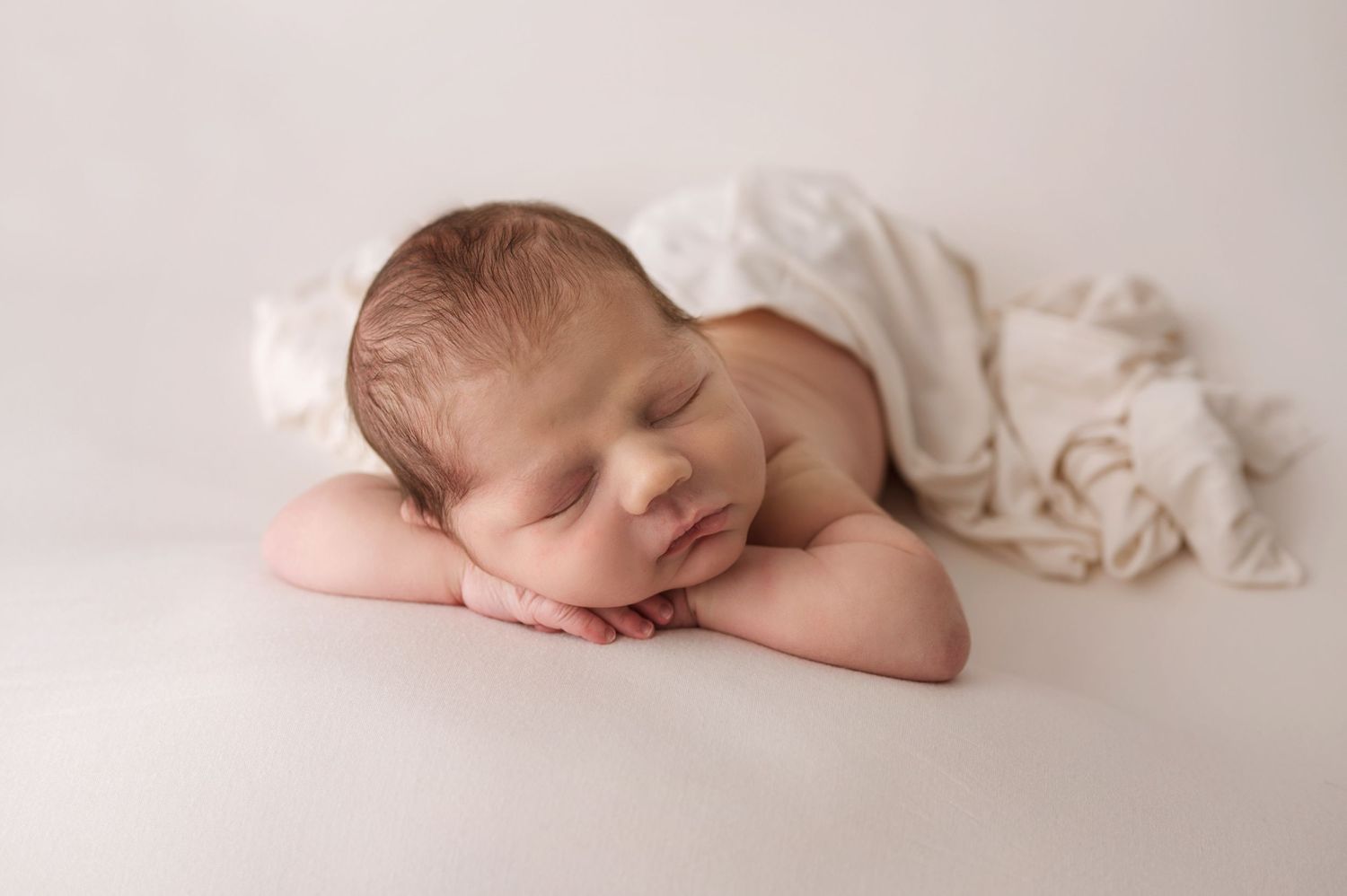 A sleeping newborn rests peacefully on their side against a soft white backdrop draped in light fabric.