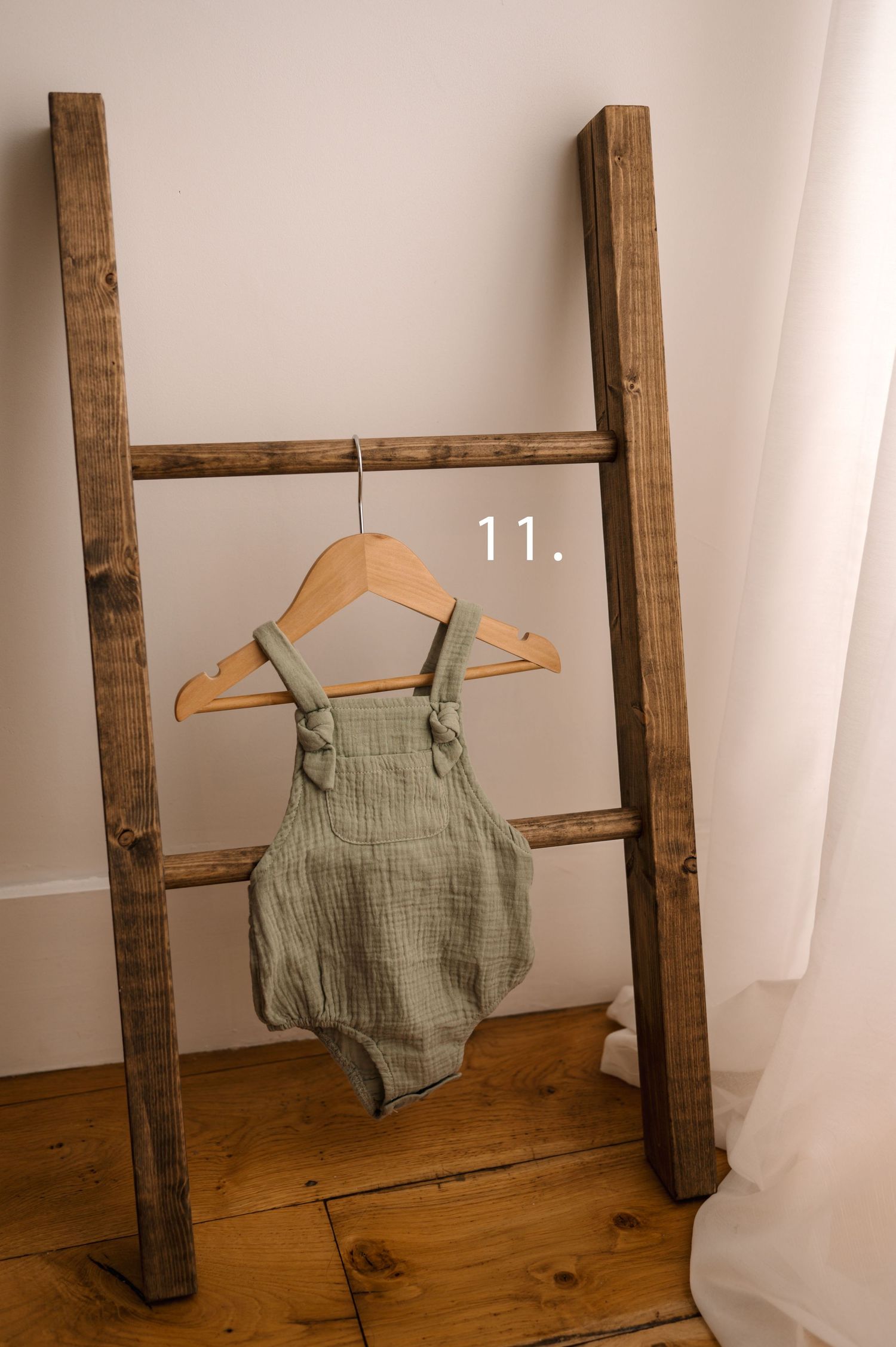 A sage green linen baby romper displayed on a wooden hanger against a white backdrop with wooden ladder decor.