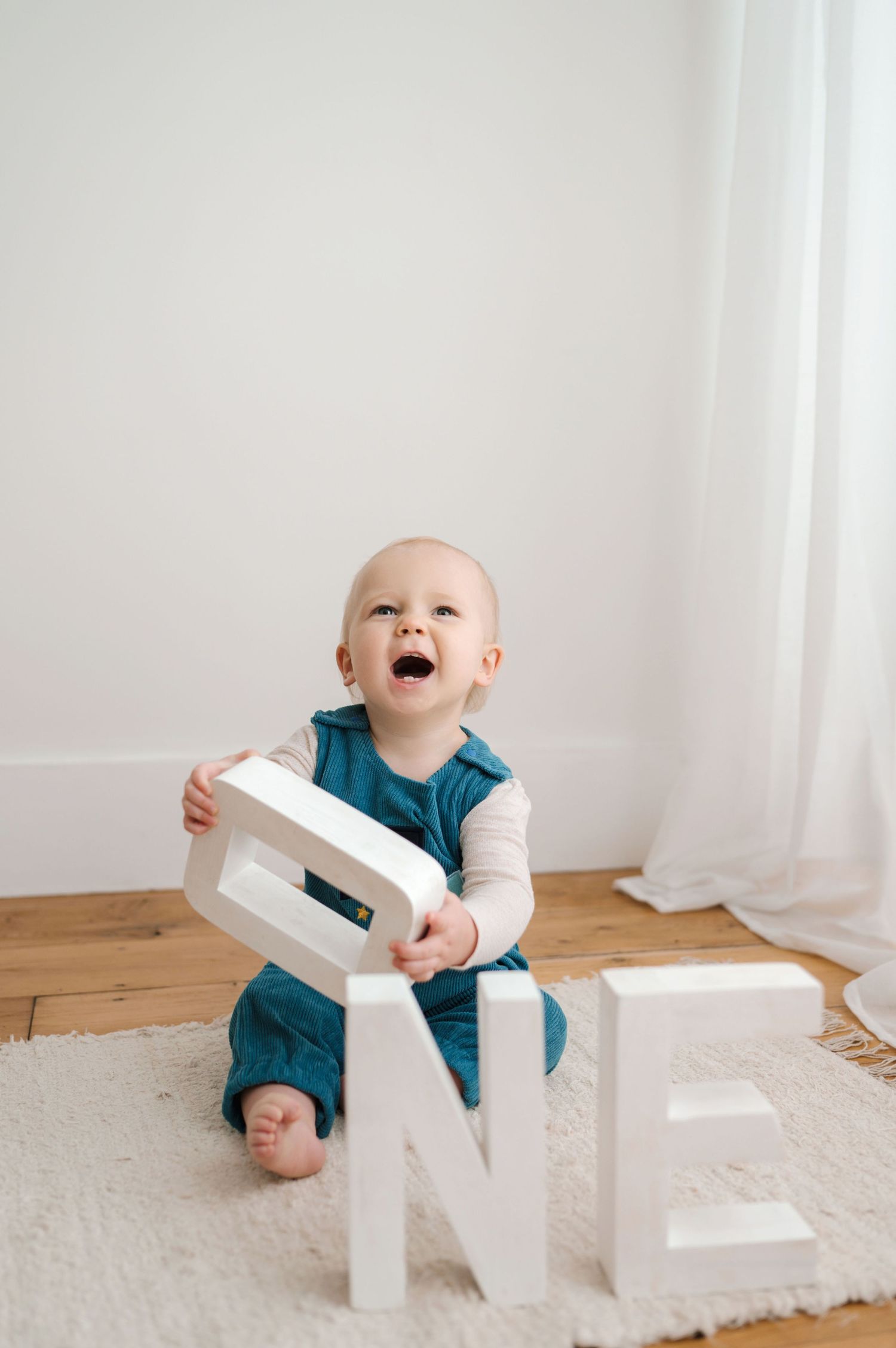 Baby in teal romper plays with white letter blocks on plush carpet while laughing and exploring.