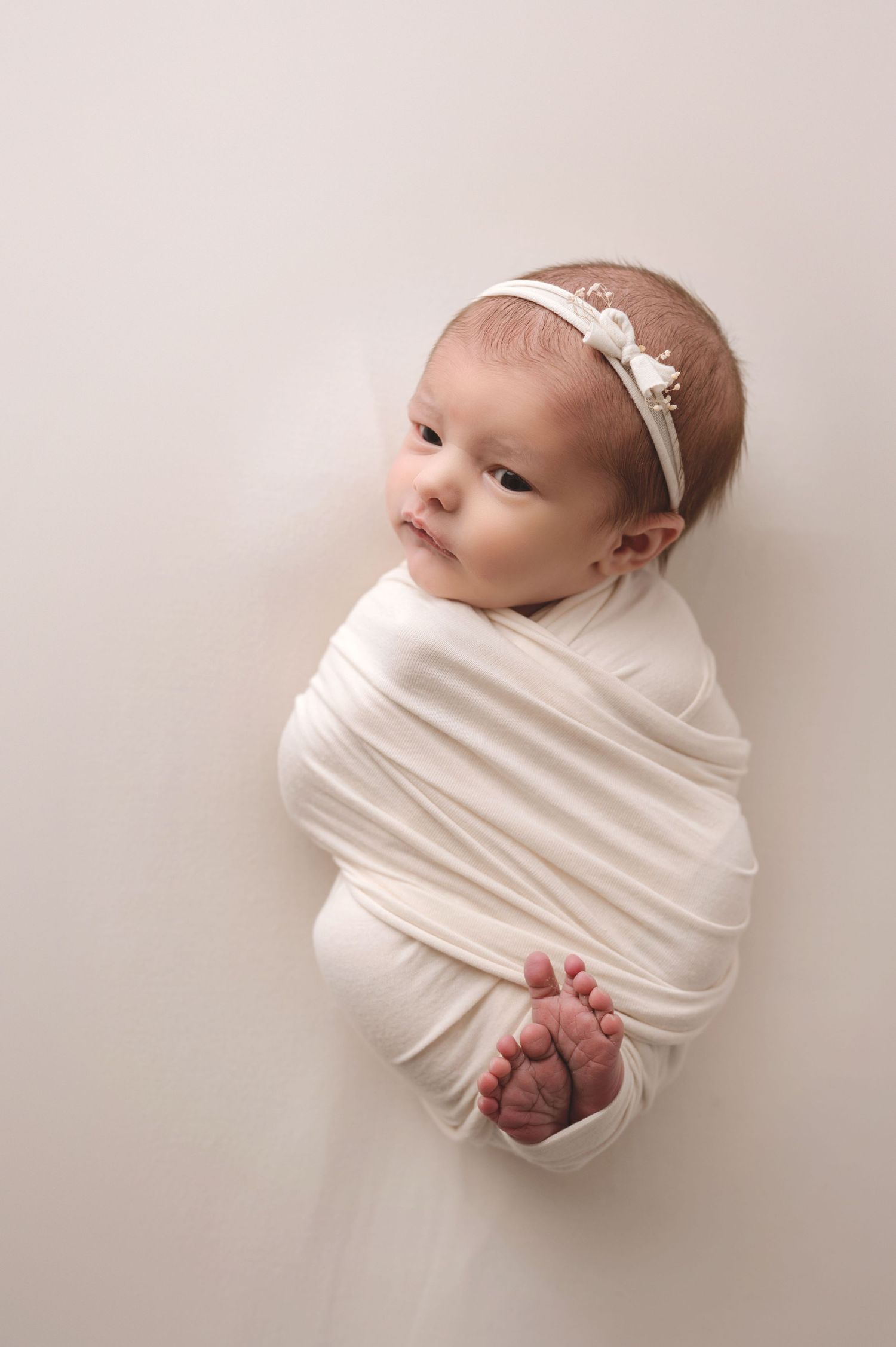 A newborn baby swaddled in white fabric wearing a delicate headband while lying on a light backdrop.