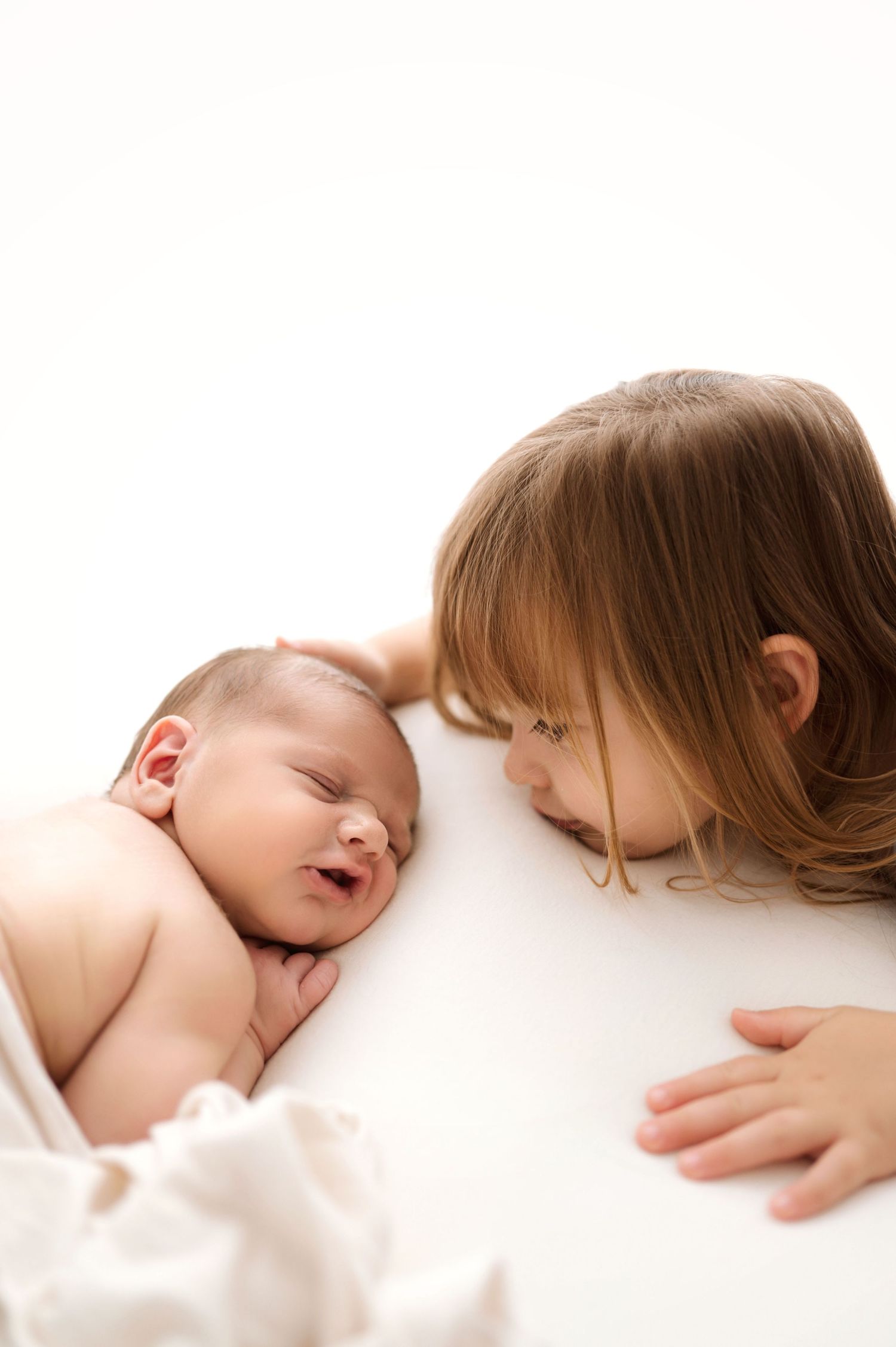 A young girl gazes lovingly at a smiling newborn baby lying on a white surface.