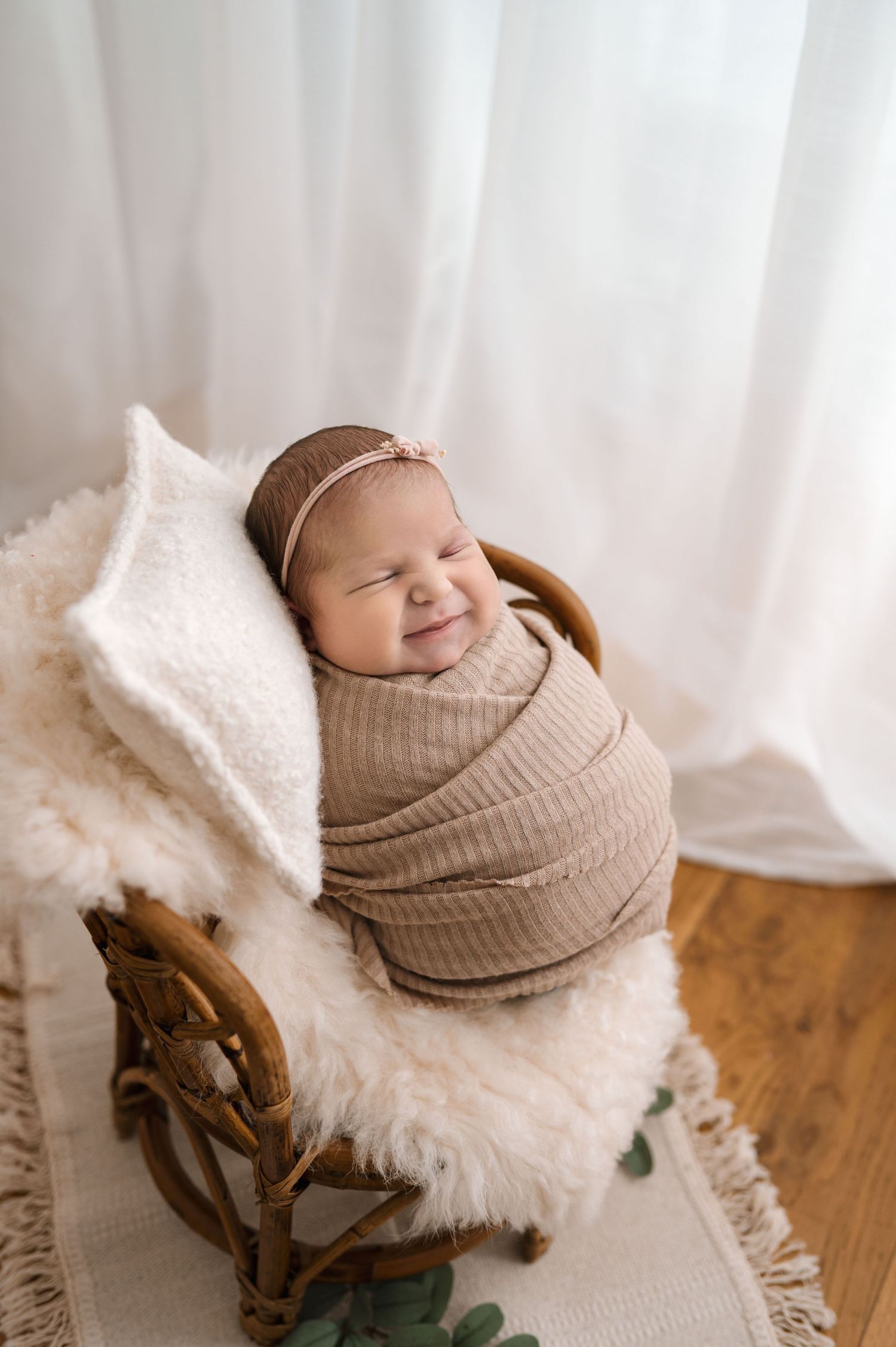 Smiling newborn baby swaddled in a tan wrap, sitting in a small wicker chair on a fluffy white rug.