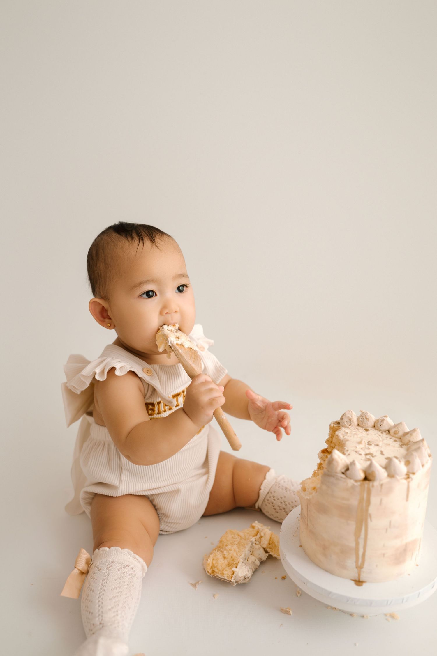Baby girl in cream romper eating cake during first birthday smash cake photo session.