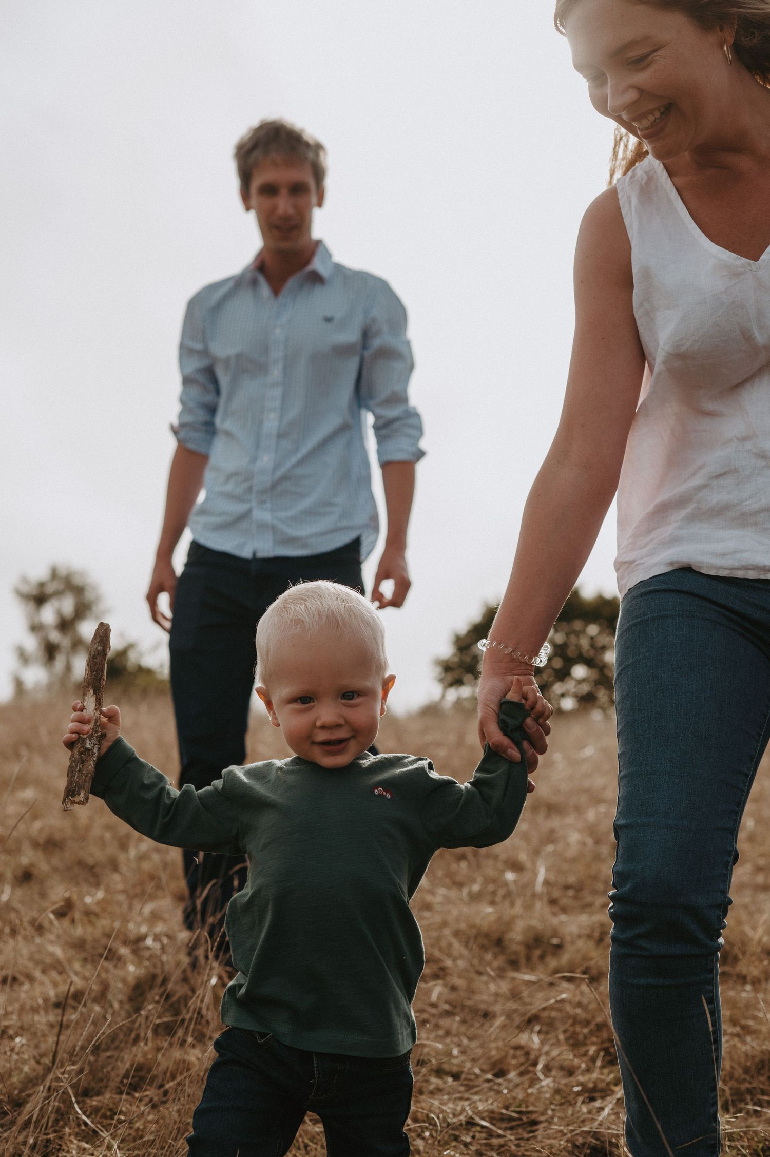 A toddler in a dark sweater walks on a field holding hands with family members while smiling.