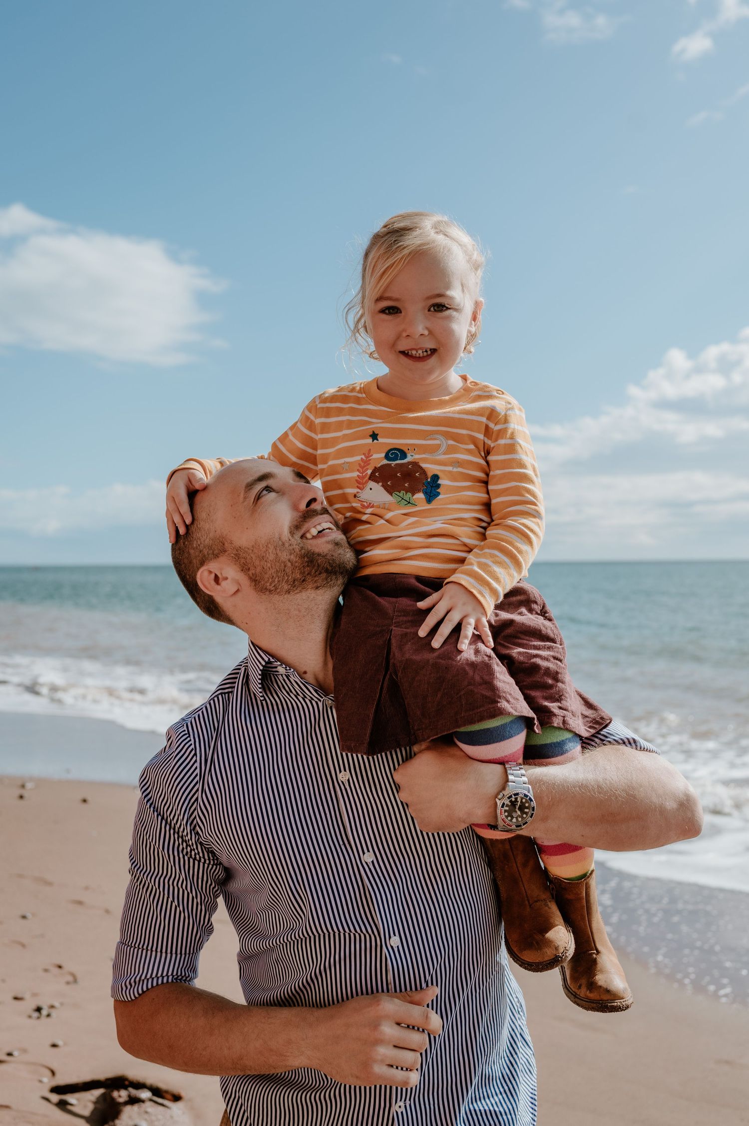 Man holding toddler on shoulders at beach, child laughing in striped shirt, sunny sky and ocean in background.