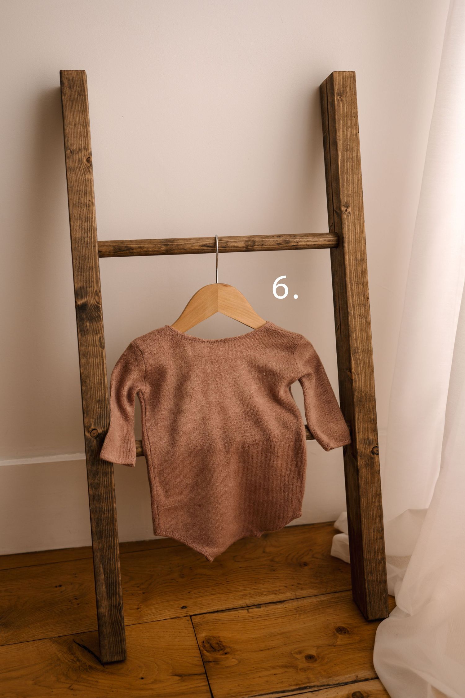 A dusty pink baby bodysuit hangs on a wooden ladder against a white wall with hardwood flooring.
