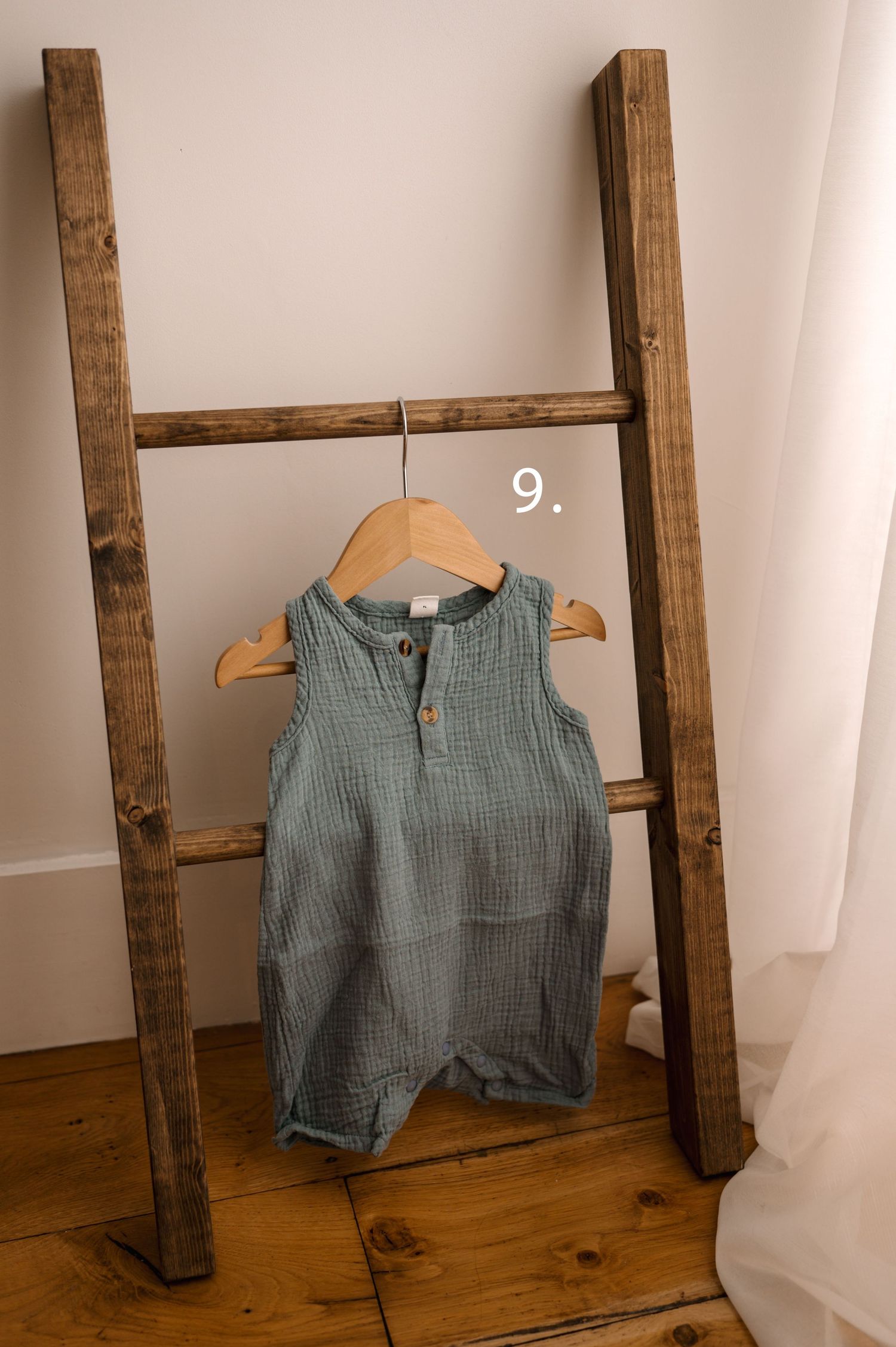 Sage green muslin baby bloomers and top set displayed on wooden hangers against a rustic ladder.