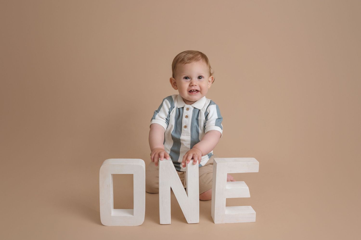 A baby in a striped outfit stands next to large white letters spelling ONE against a neutral beige backdrop.