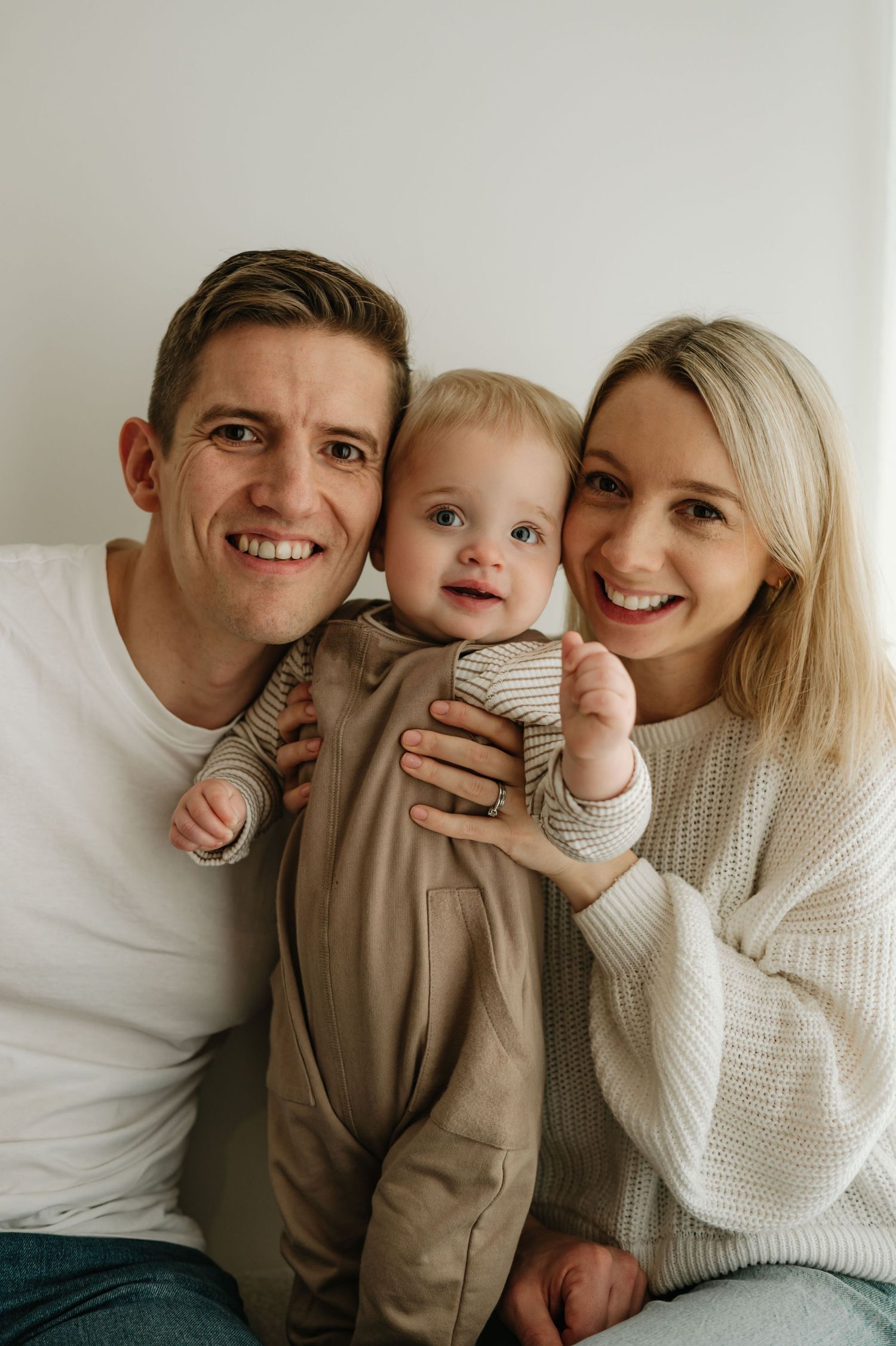 Happy young family portrait with smiling parents and adorable baby in neutral tones against white background.
