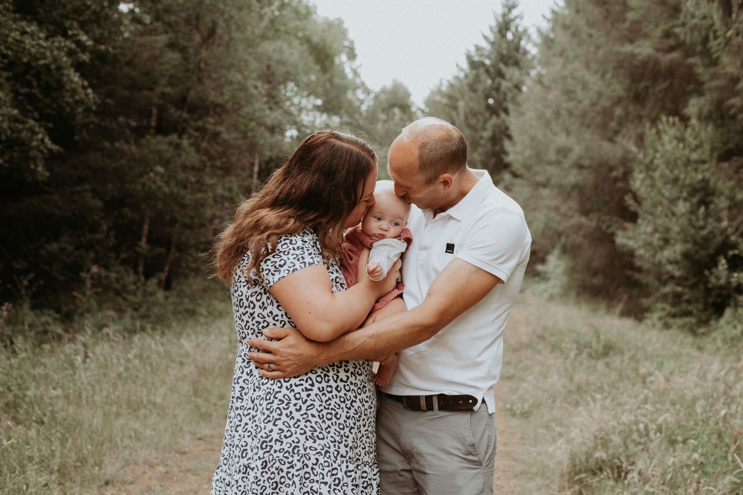 A couple shares a tender embrace while standing in a sunlit forest clearing among pine trees with their baby