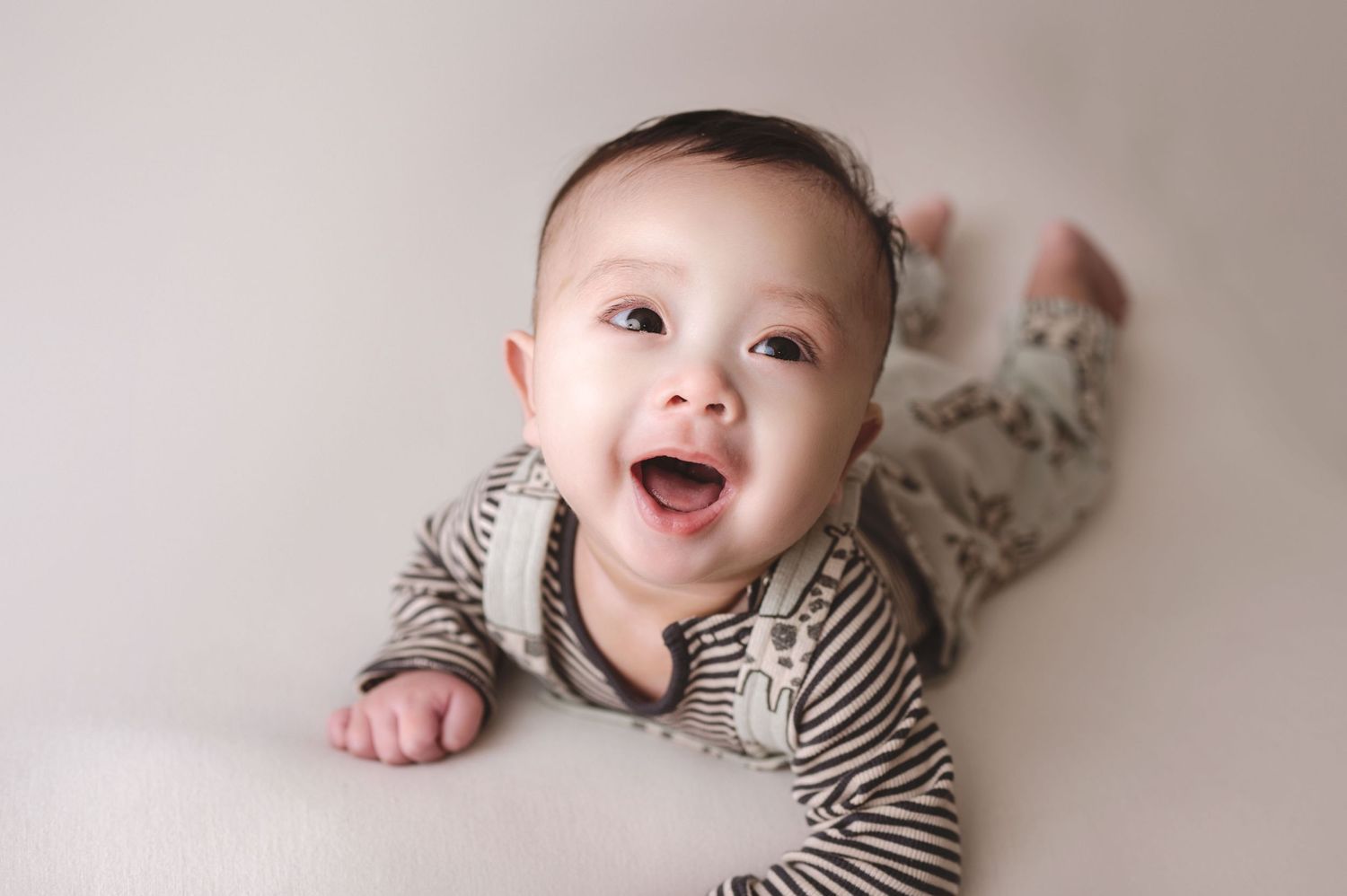A joyful baby in a striped onesie lies on their tummy on a light gray background, smiling widely with delight.