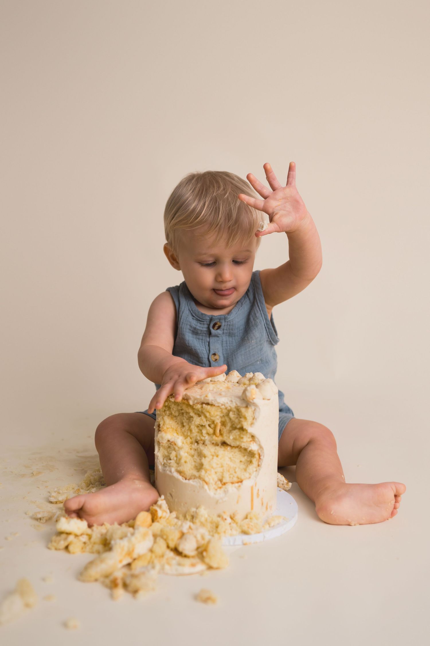 Baby in denim romper smashes and plays with a messy white cake during cake smash photo session.