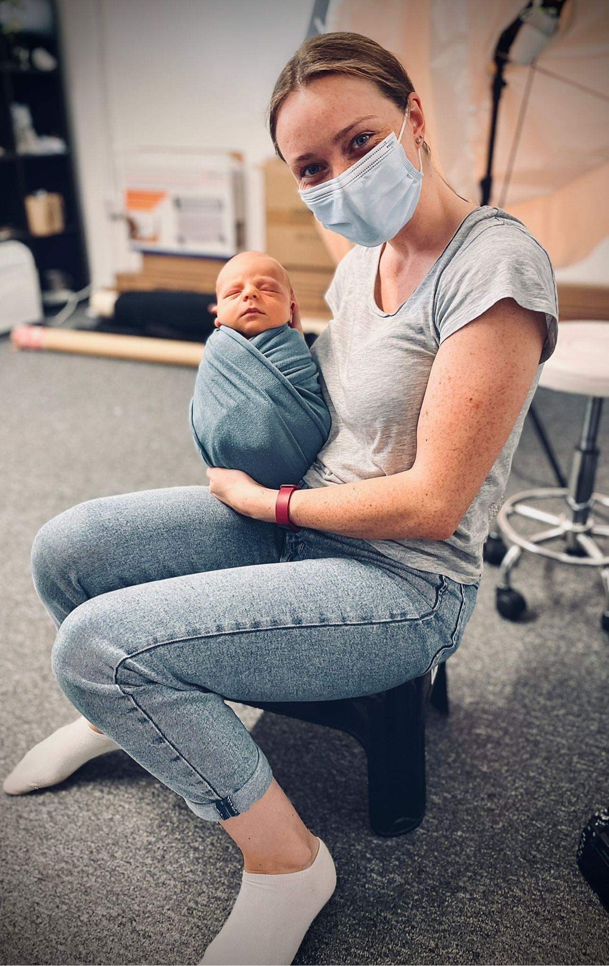 Pengelly Portraits wearing protective face mask holds sleeping newborn baby wrapped in blue swaddle in photo studio.