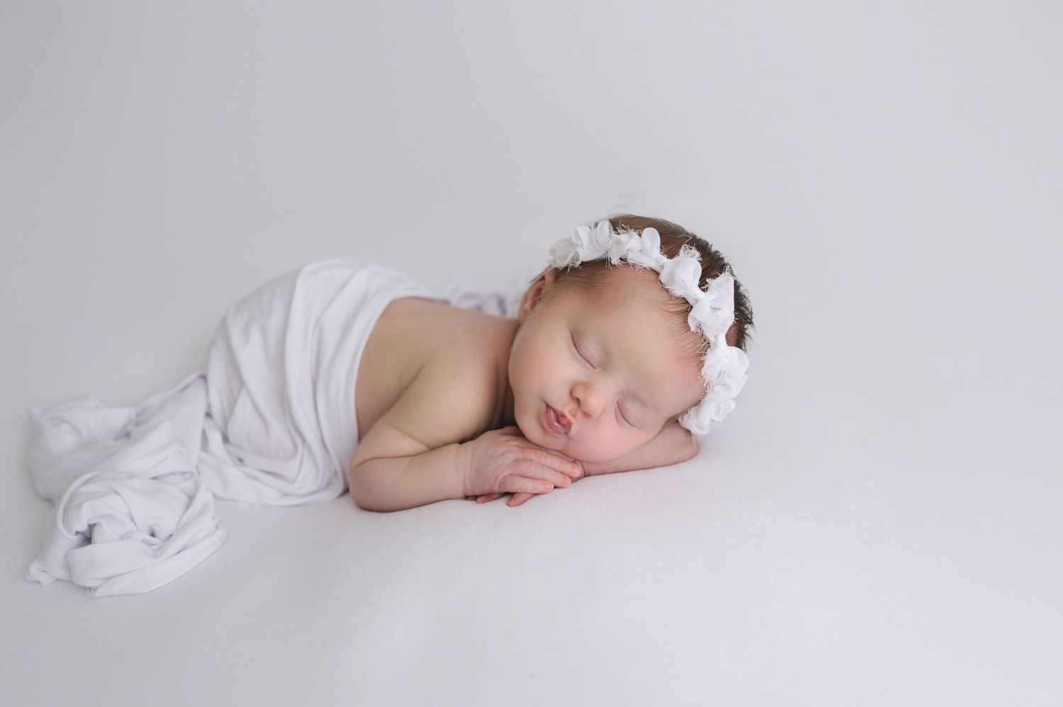 Sleeping newborn baby wrapped in white cloth with floral headband on white background.