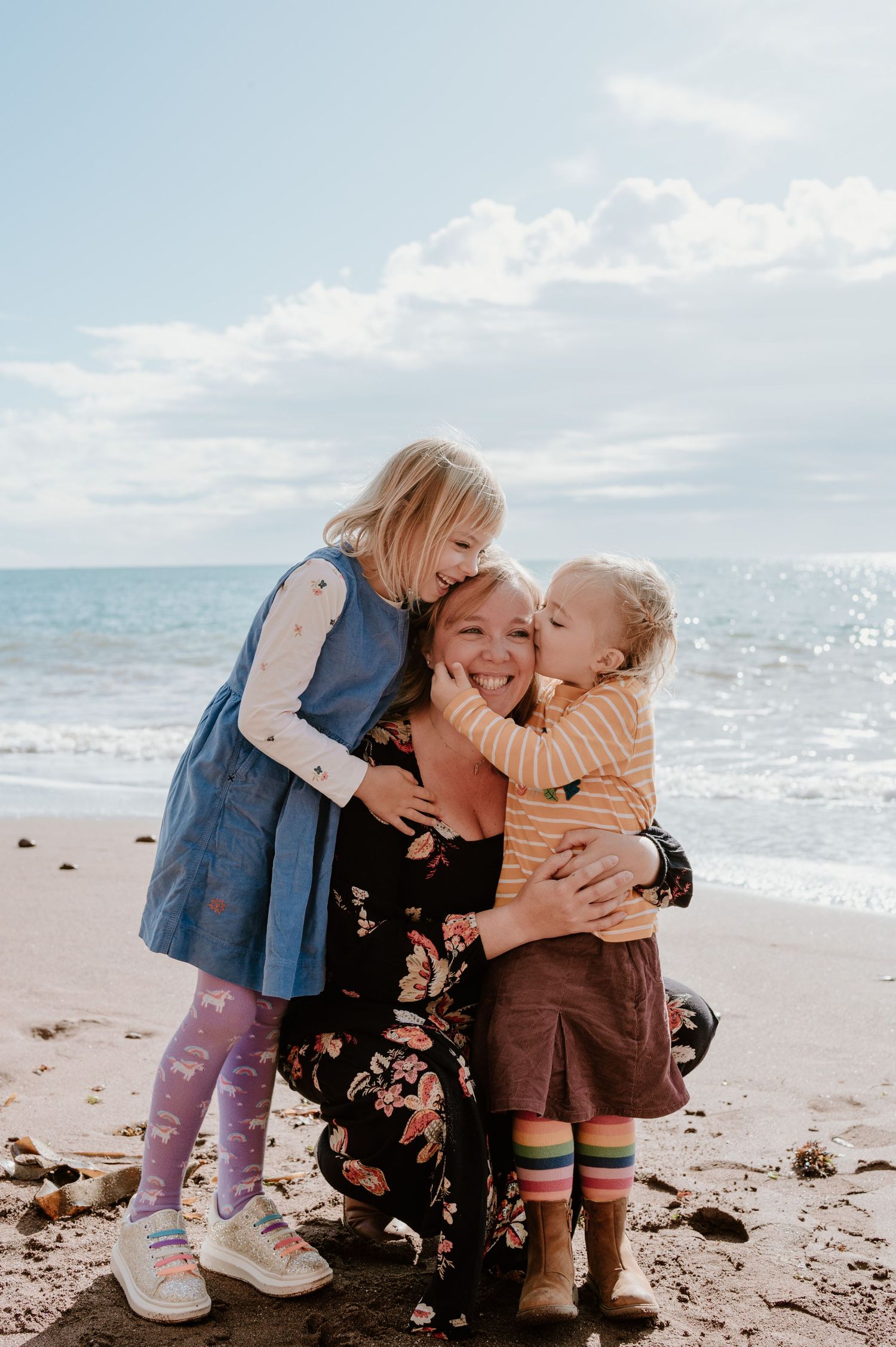 A mother crouches on a sandy beach while her two young blonde daughters hug and kiss her cheeks, all smiling joyfully.