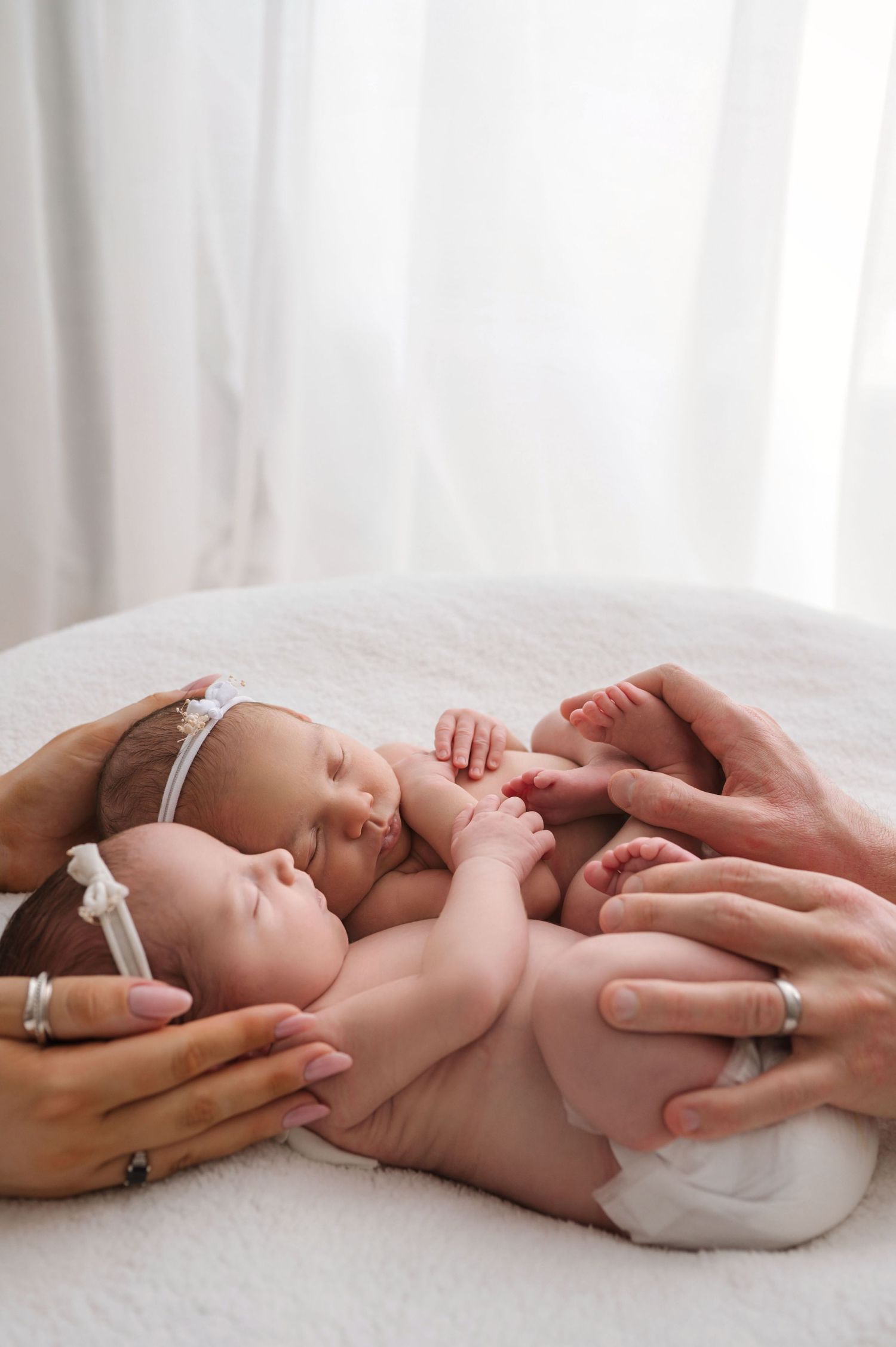 Two newborns lie together on a white surface while gentle hands tenderly touch their tiny bodies.