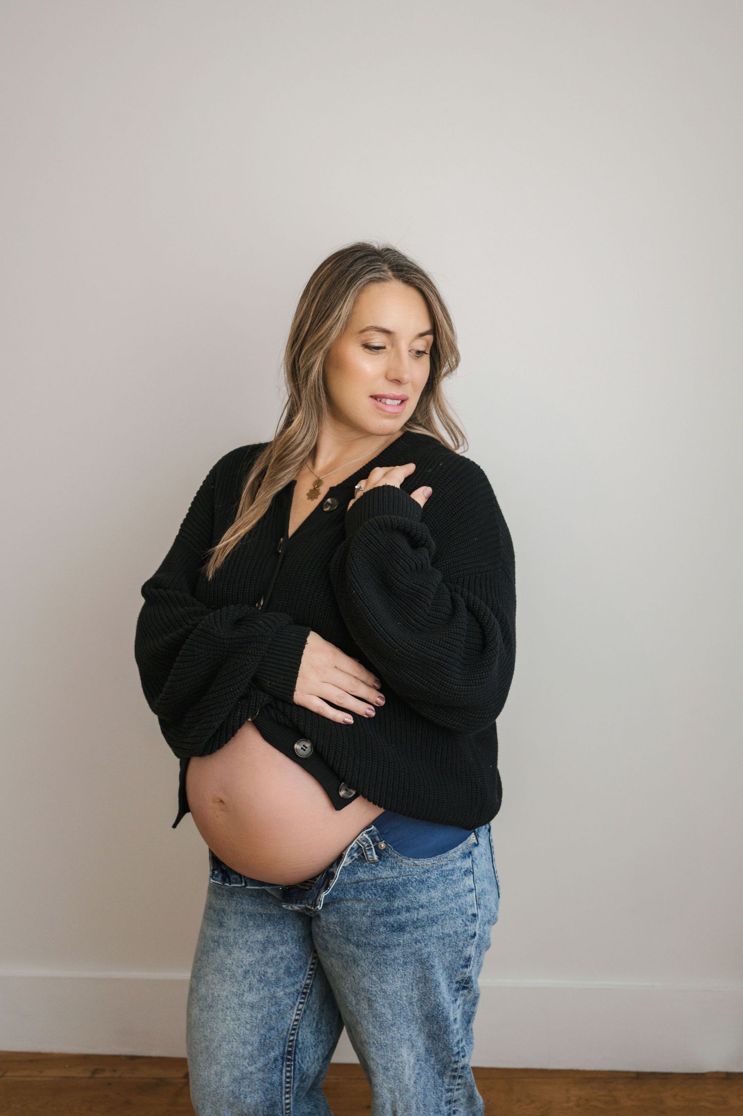 Pregnant woman in black cardigan and jeans cradling her bare baby bump during maternity photoshoot.