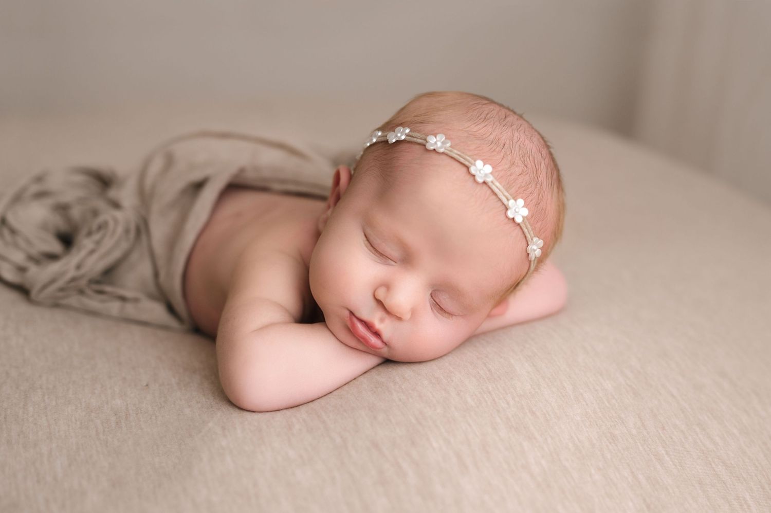 A sleeping newborn baby wrapped in soft beige fabric lies peacefully on a neutral backdrop during a professional photoshoot.