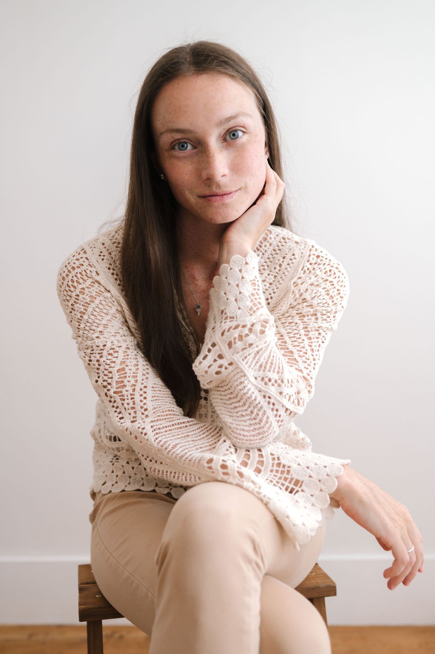 A person in a cream knit sweater and beige pants sits casually on a wooden chair against a white wall.
