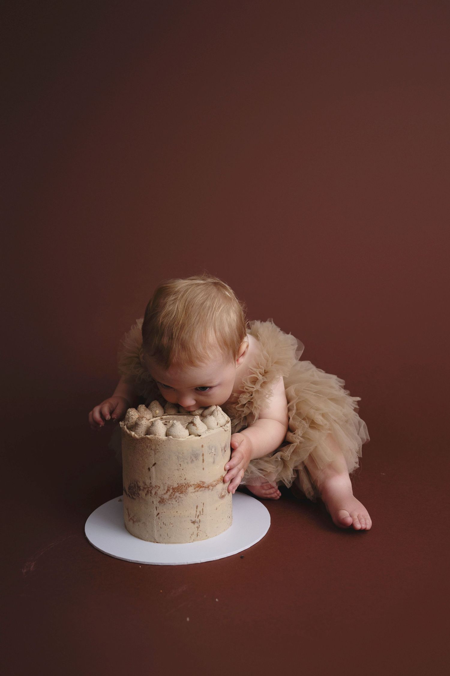 babyA small figure in a tulle dress leans over to taste a rustic brown frosted cake on a white plate against dark backdrop.