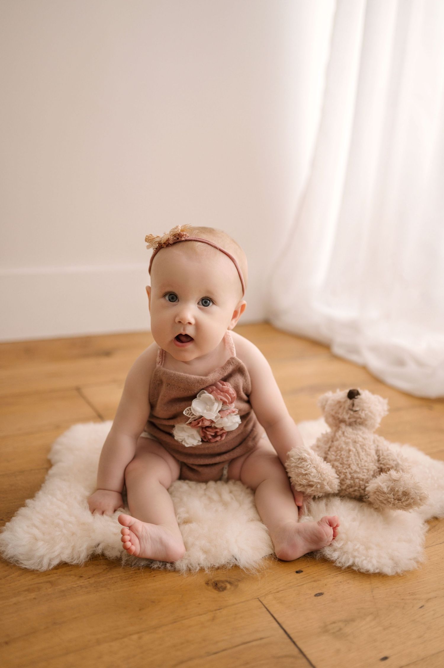 Baby wearing brown outfit sits on white fluffy rug with teddy bear on wooden floor in natural light setting.
