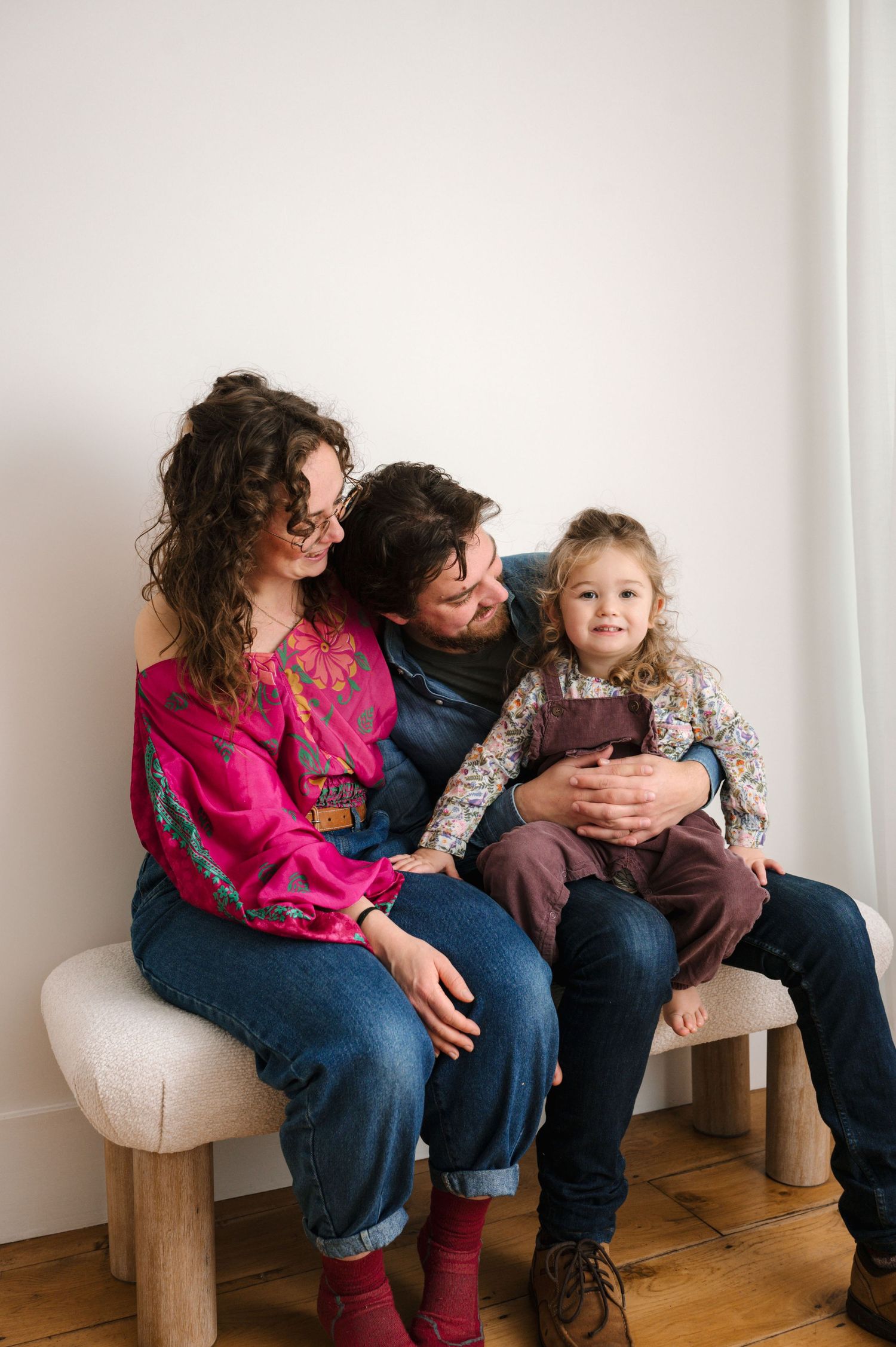 A family sits together on a white bench against a neutral wall, wearing casual clothes in bright colors and denim.