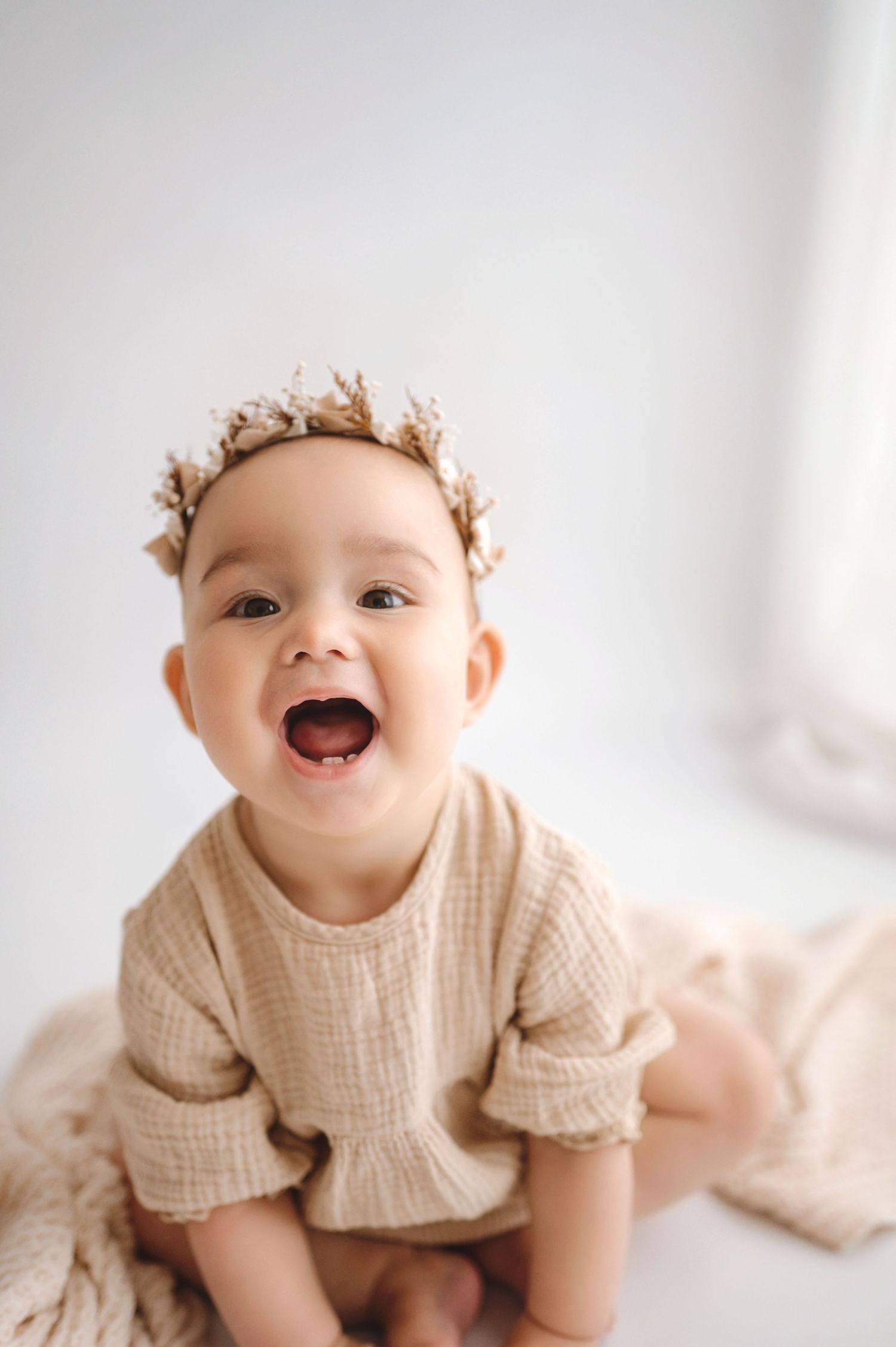 Adorable baby girl in beige muslin outfit and floral crown smiling joyfully in a bright white studio.