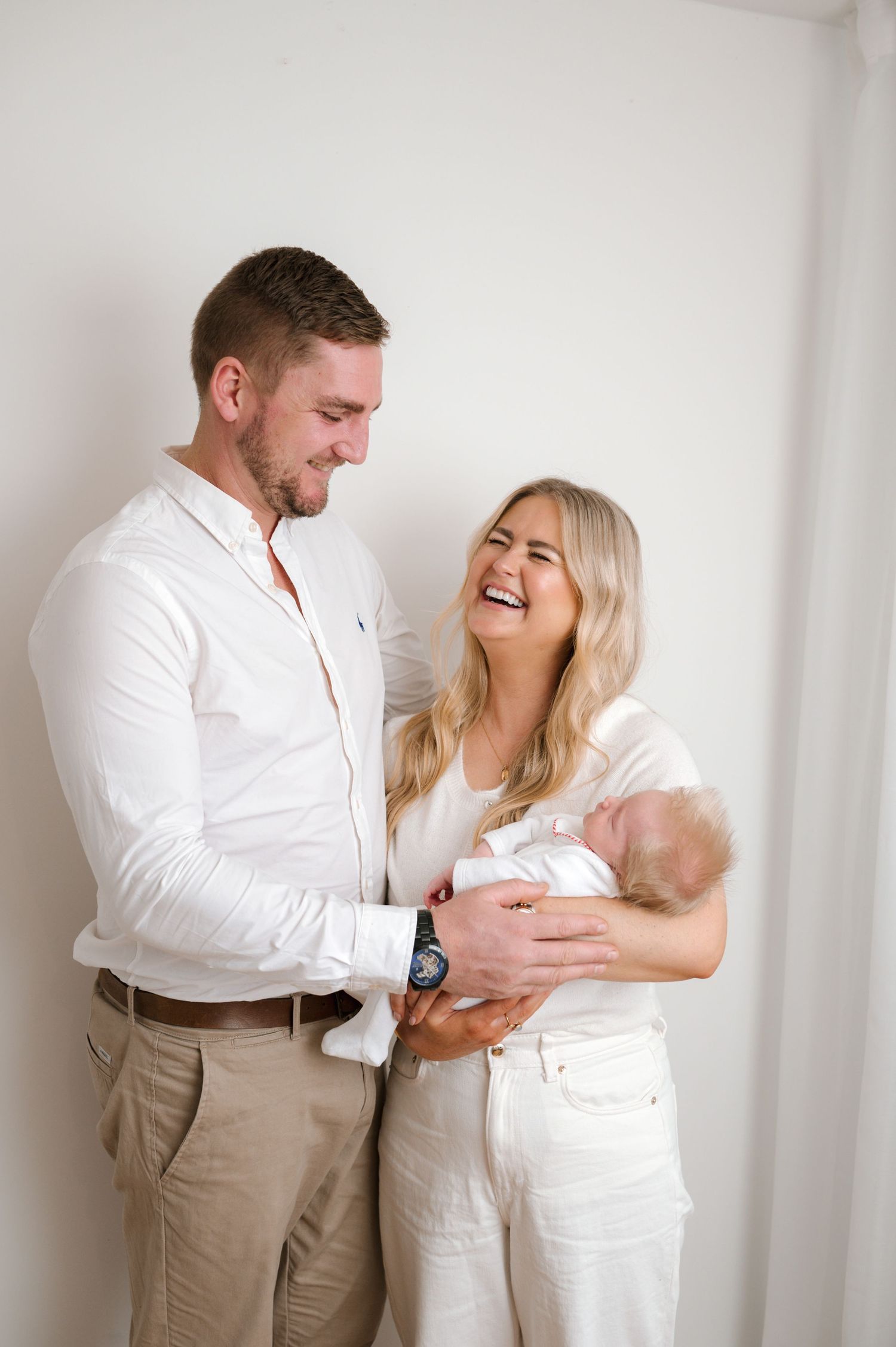 Happy couple in white outfits smiling while holding their newborn baby against a light background.