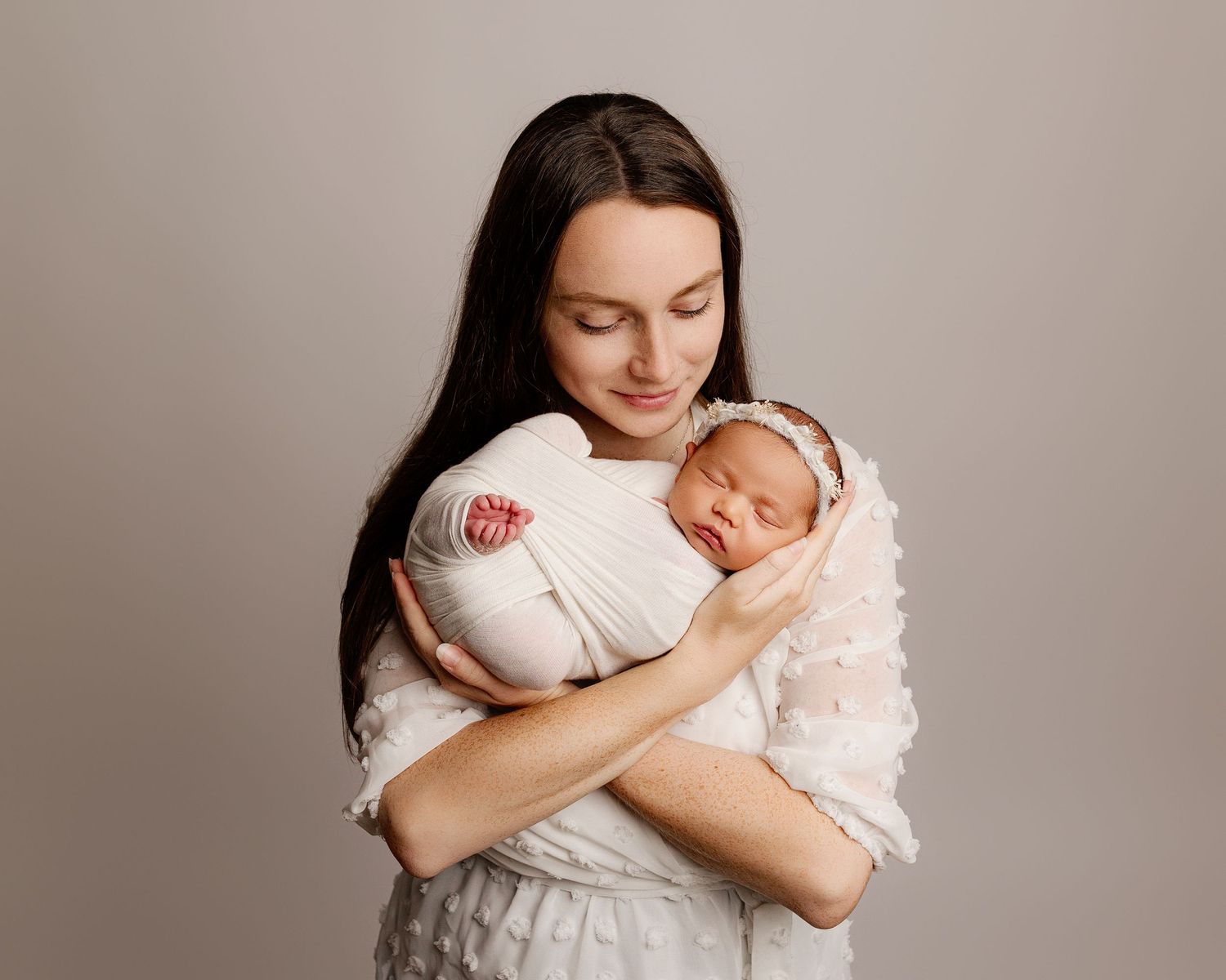 A mother in a white dress cradles her sleeping newborn baby wrapped in white against a neutral background.