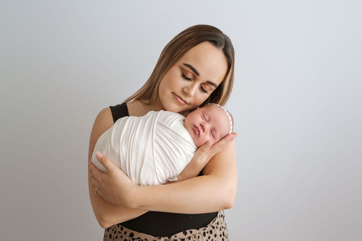A young mother tenderly holds her swaddled newborn baby close to her chest against a soft gray background.